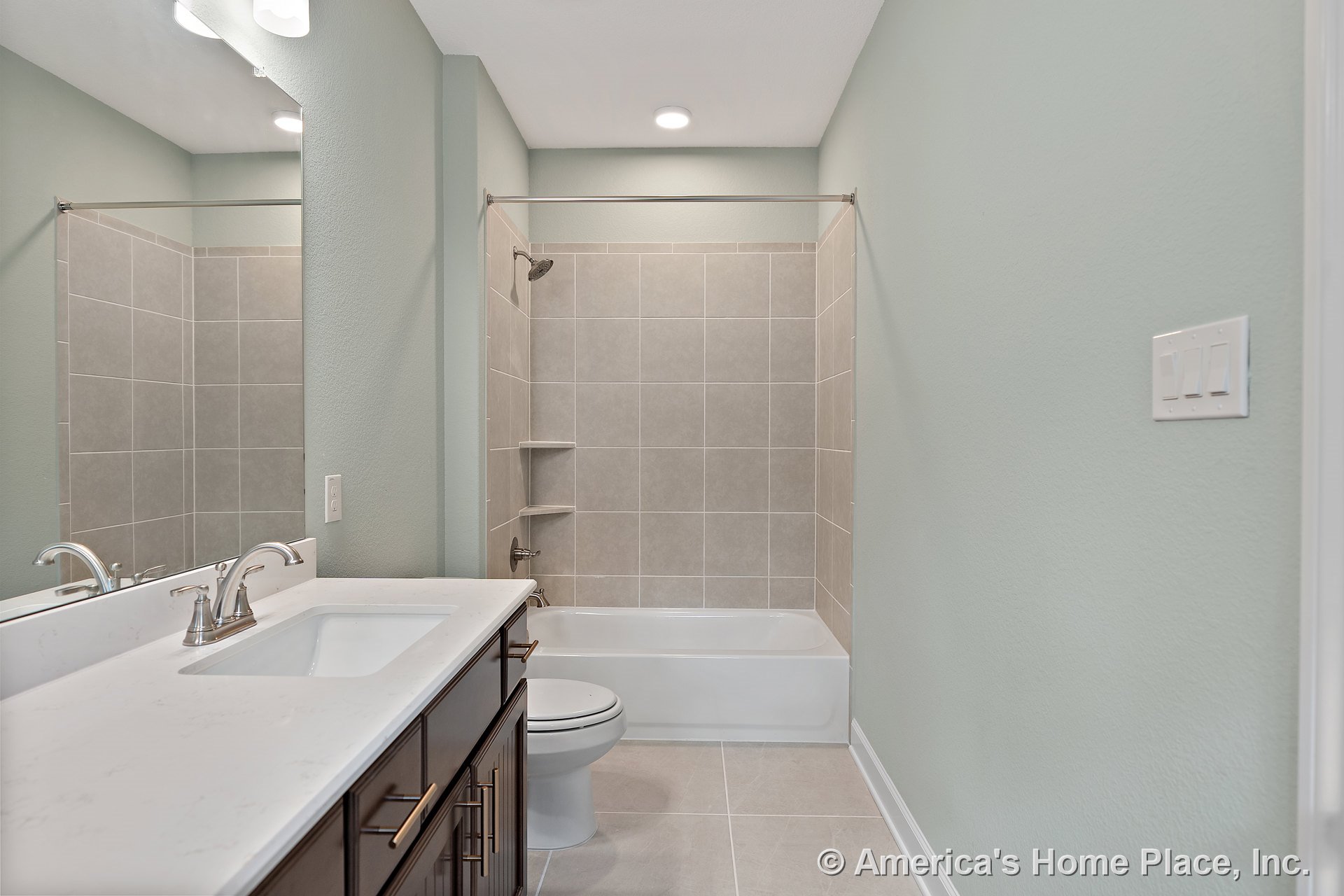 Tiled shower-tub combo with built-in shelves, quartz countertop on dark wood vanity, rectangular undermount sink, brushed nickel faucet, large wall mirror, recessed ceiling light