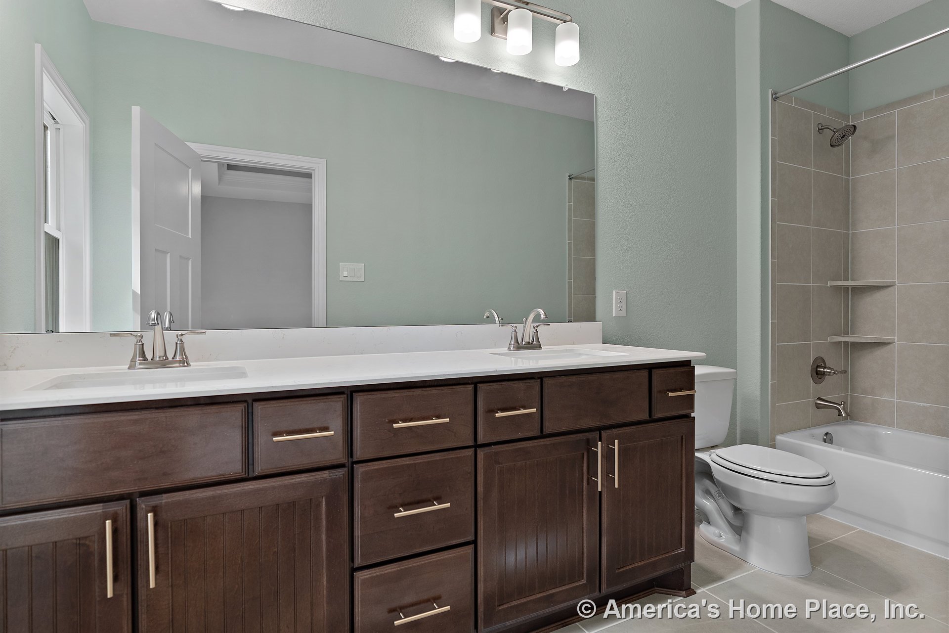 Double vanity with dark wood cabinets, chrome faucets, and built-in lighting beneath a large wall mirror; tile flooring extends to a bathtub with tiled surround, adjacent toilet