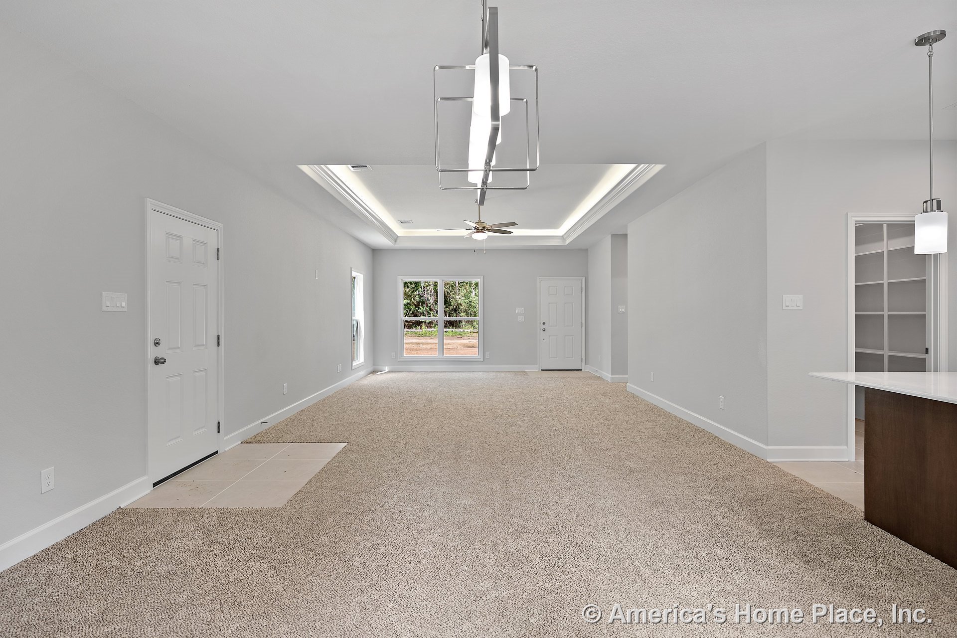 Tray ceiling with recessed lighting and modern ceiling fan above carpeted living area; large double windows provide backyard views; white interior doors and built-in pantry