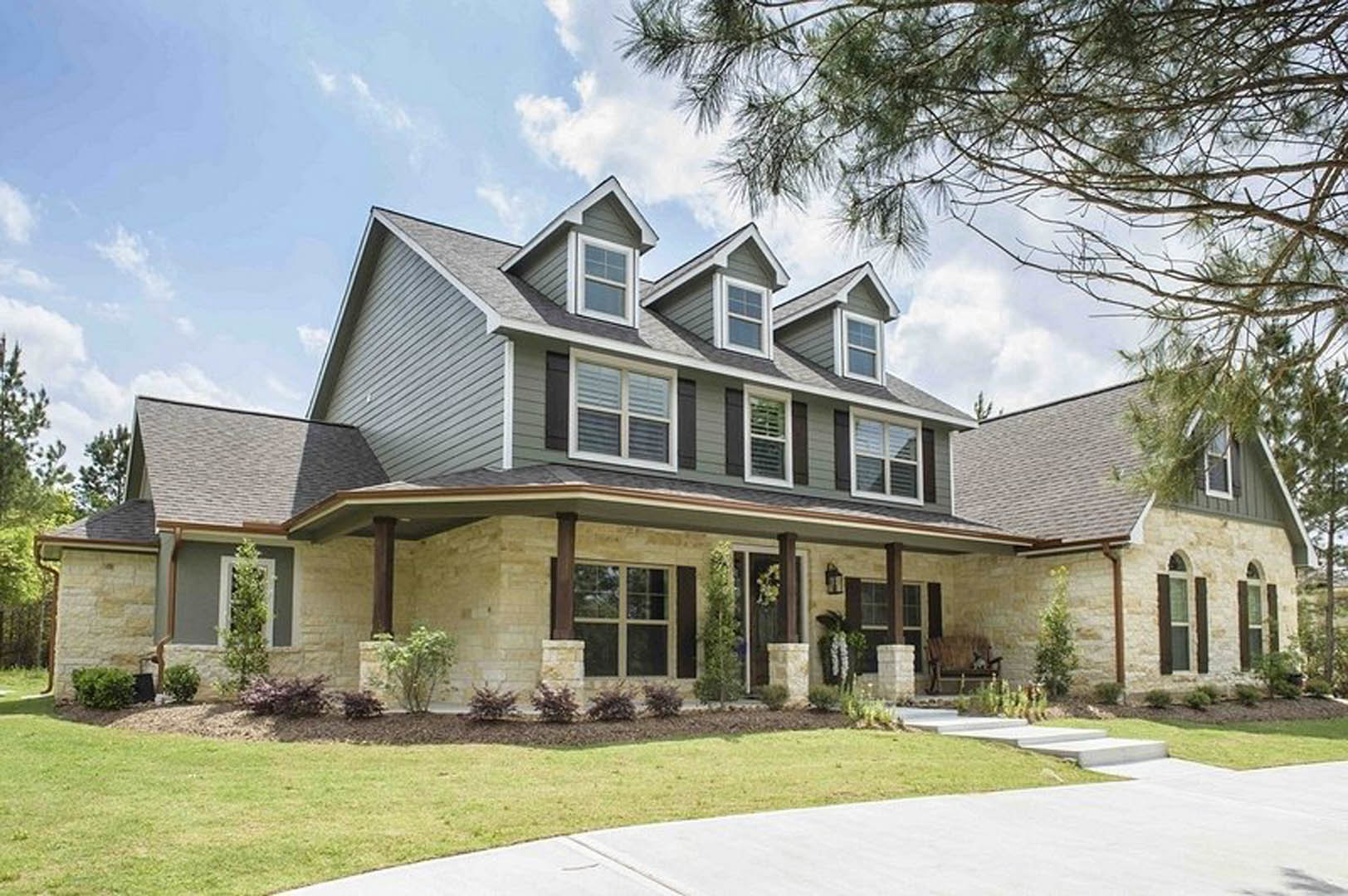 Two-story house with white-framed windows, covered front porch, concrete driveway, green lawn, and mature trees under a partly cloudy sky