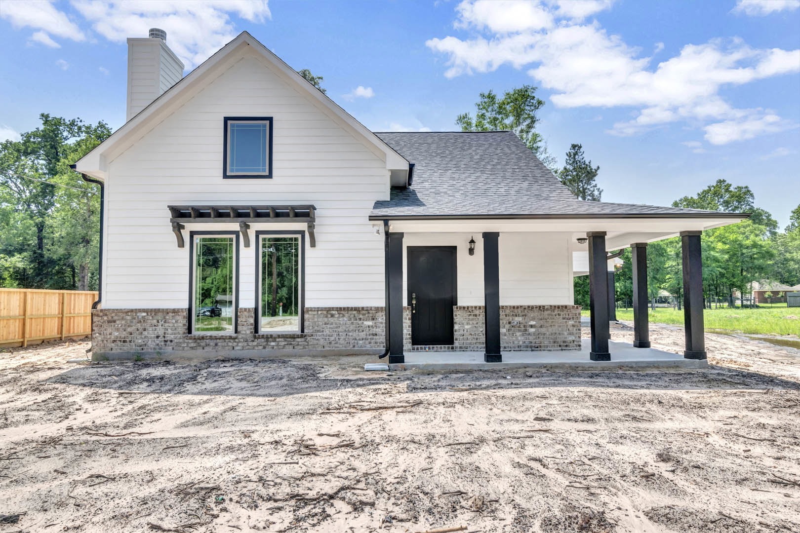 Blue and white window on a custom home exterior, black door with white trim, wooden fence in foreground, dirt road and ground with scattered sticks, tree visible through window