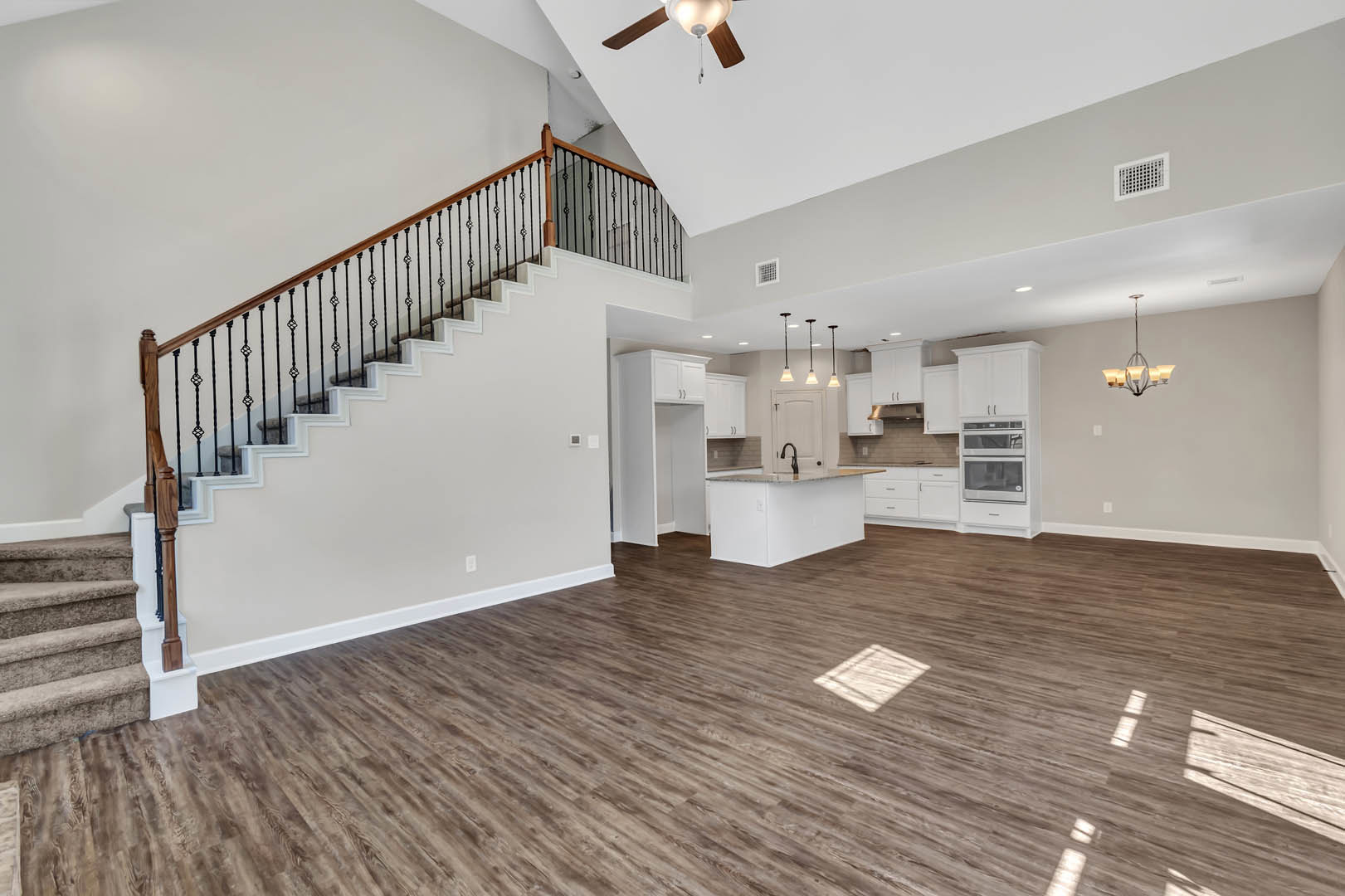 Spacious open-concept living area featuring wood flooring, a central staircase with white handrail, modern kitchen with stainless steel appliances, white vent on wall, and plaster