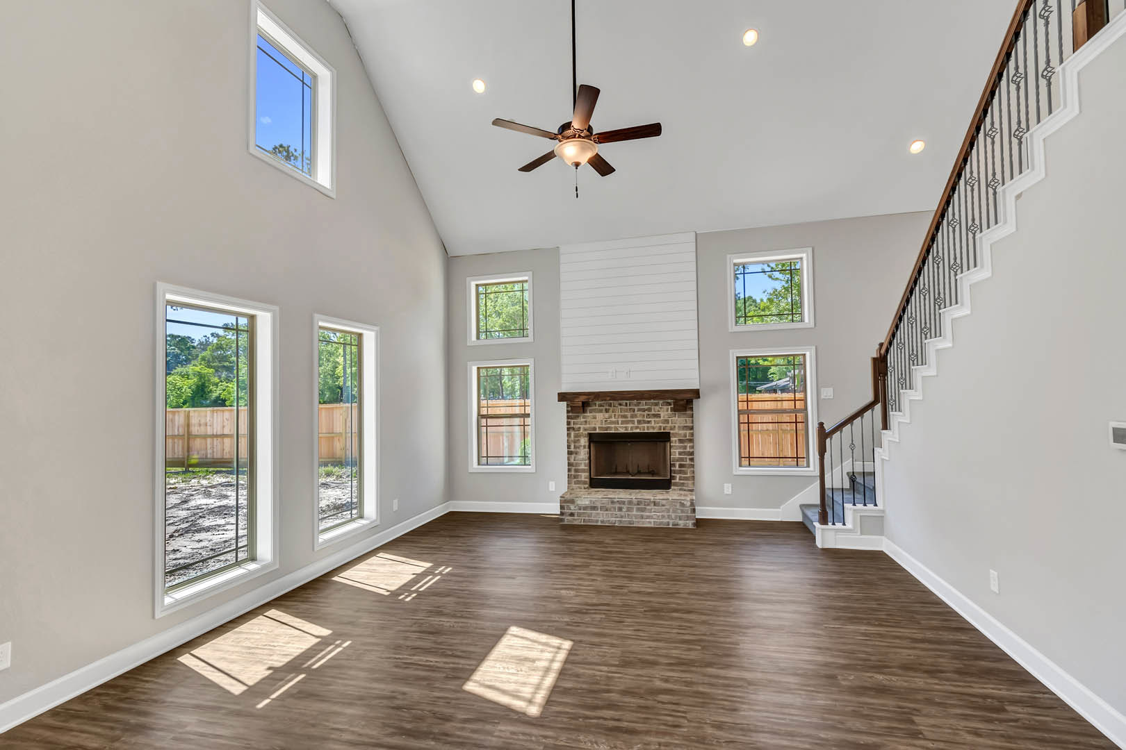 Living room with brick fireplace, wood mantel shelf, wooden flooring, ceiling fan with light, staircase, and window.