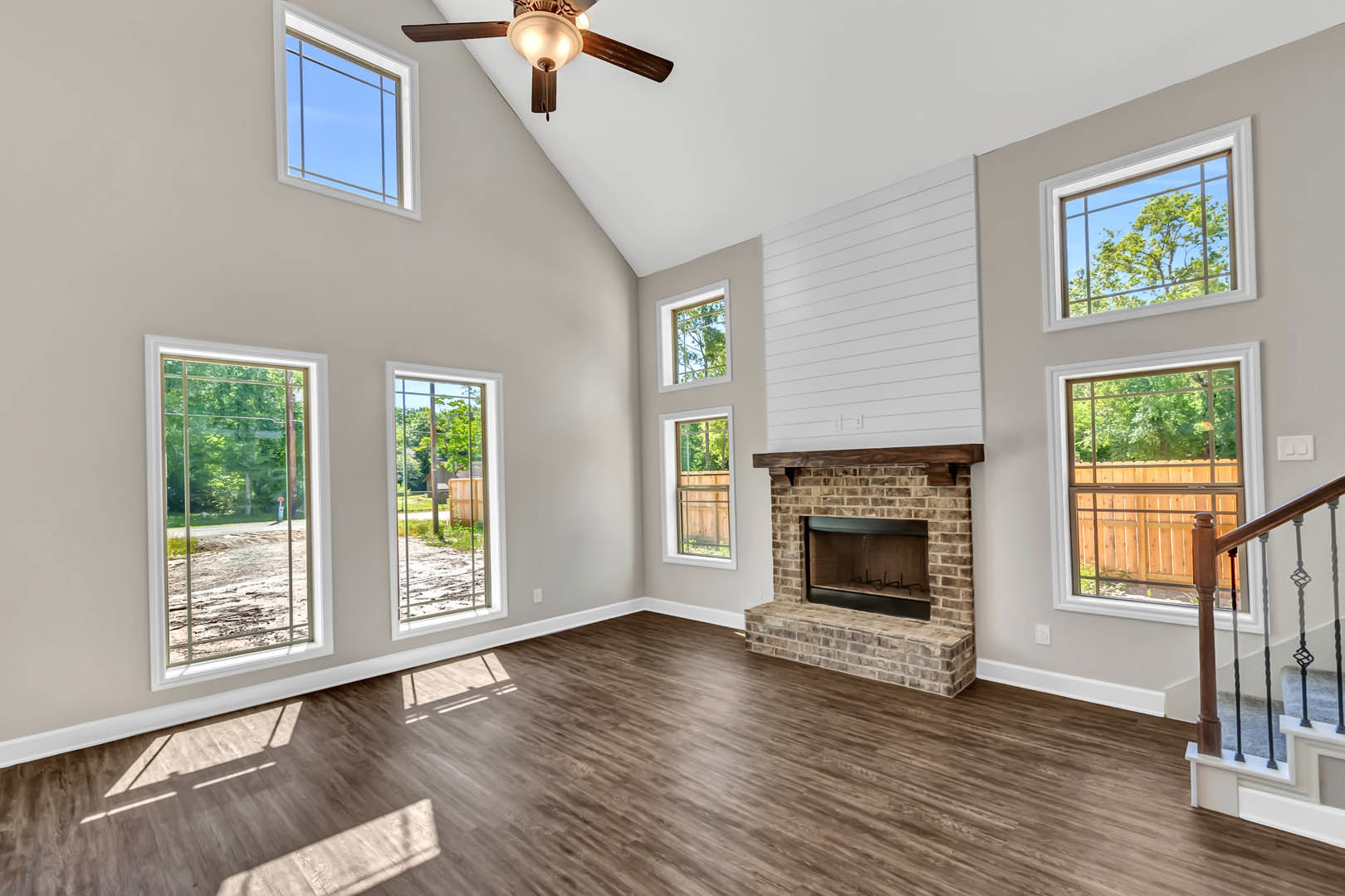 Living room with wood flooring, black-framed fireplace, large windows overlooking road and trees, white walls, ceiling fan with light, and metal railing