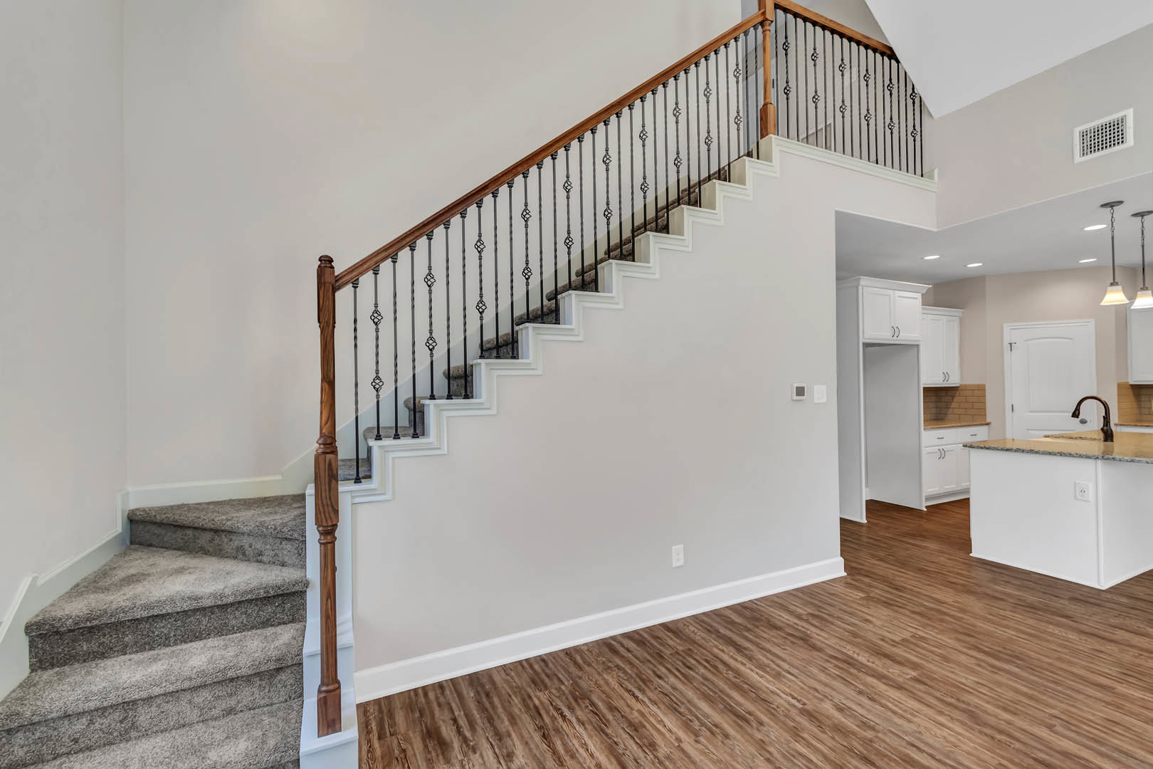 Wood staircase with white balusters and handrail, marble countertop in kitchen, wood flooring, white vent on wall, white door, modern indoor finishes
