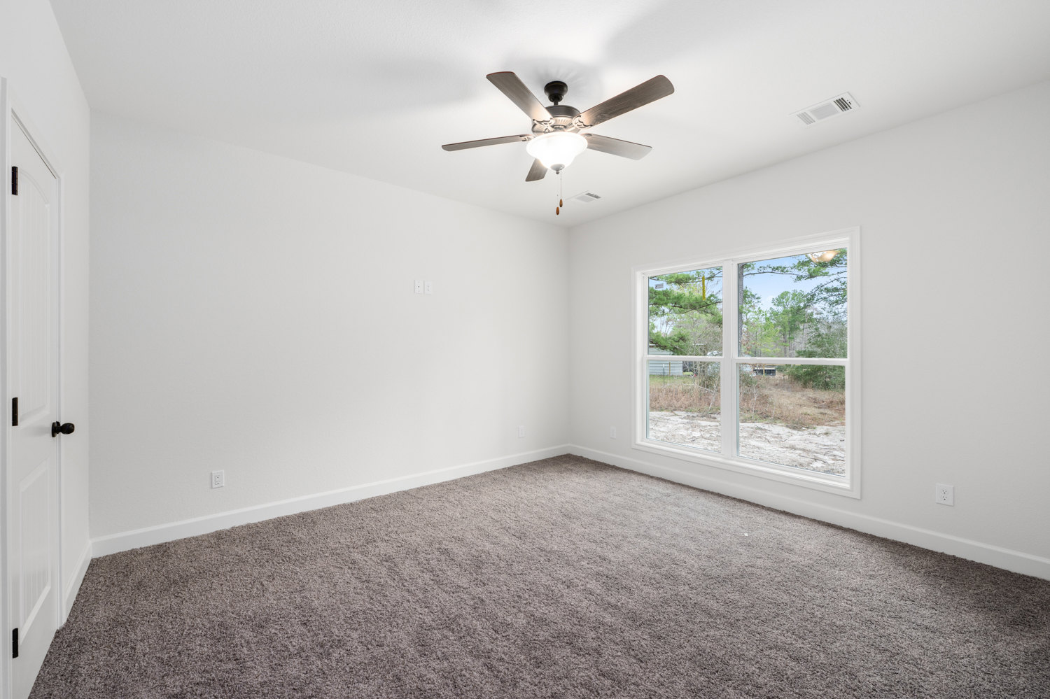 Neutral-toned carpeted room with a ceiling fan and light fixture, white walls, window showing leafy trees, and a white door with black hardware