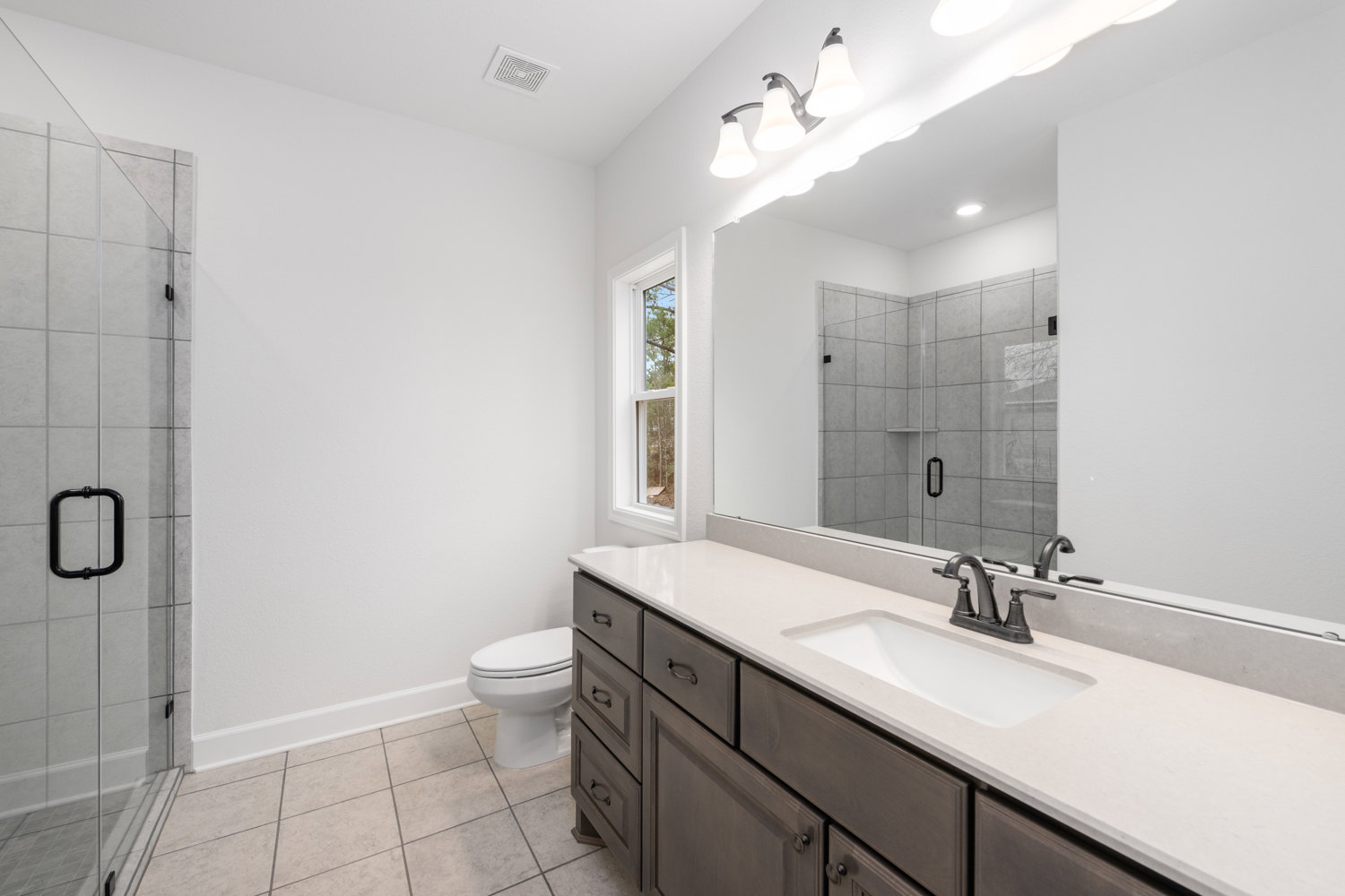 Bathroom with a wide frameless mirror above a white countertop sink, black metal faucet and handle accents, light gray tile walls, and a modern toilet visible in the background.