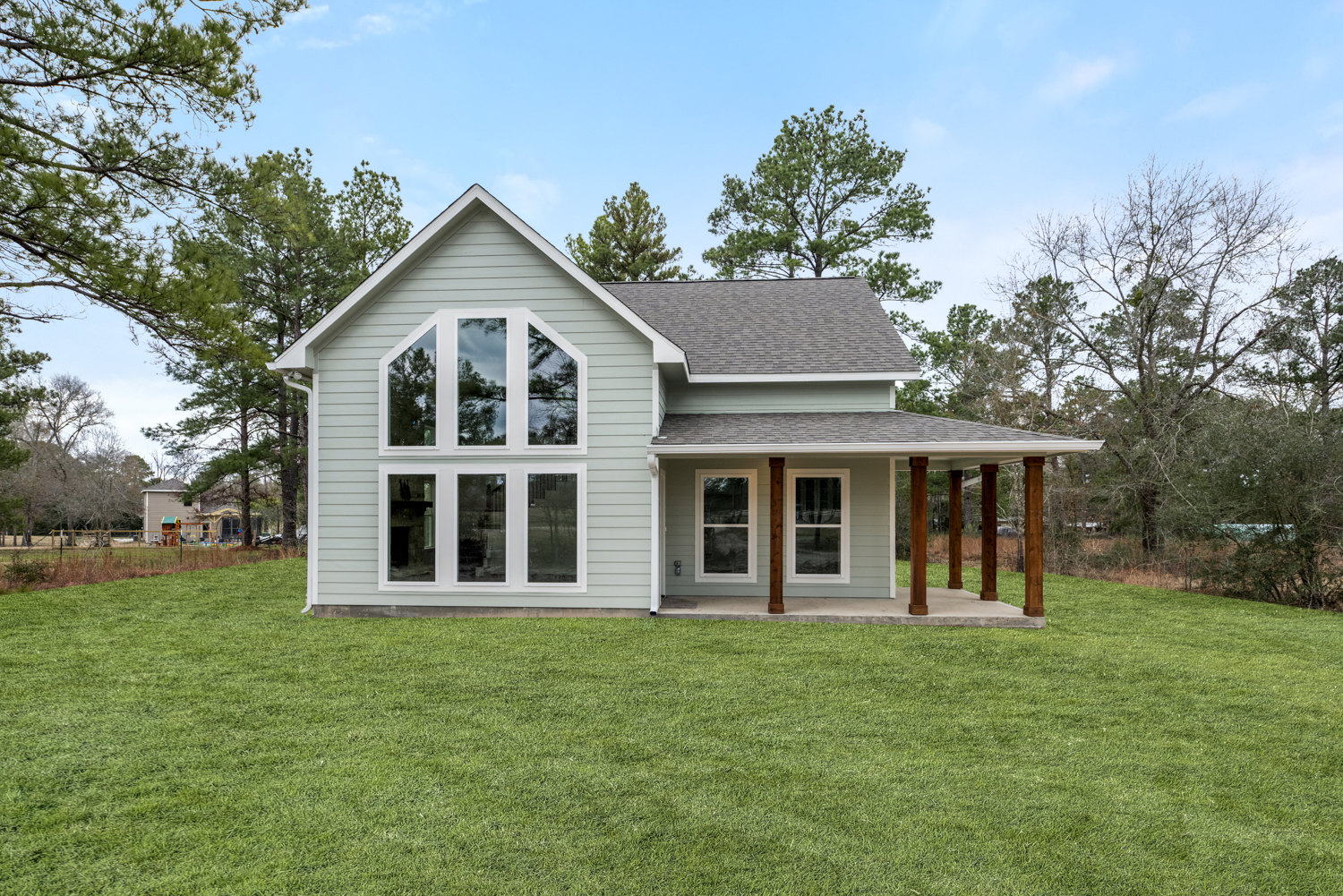 Two-story house with gray siding, large windows, covered front porch, and manicured grass lawn; mature trees and blue sky in the background.