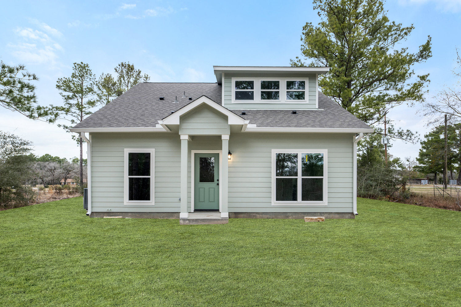 Green front door with glass window, white-framed windows, manicured lawn, and mature tree in front of a light-colored residential home.