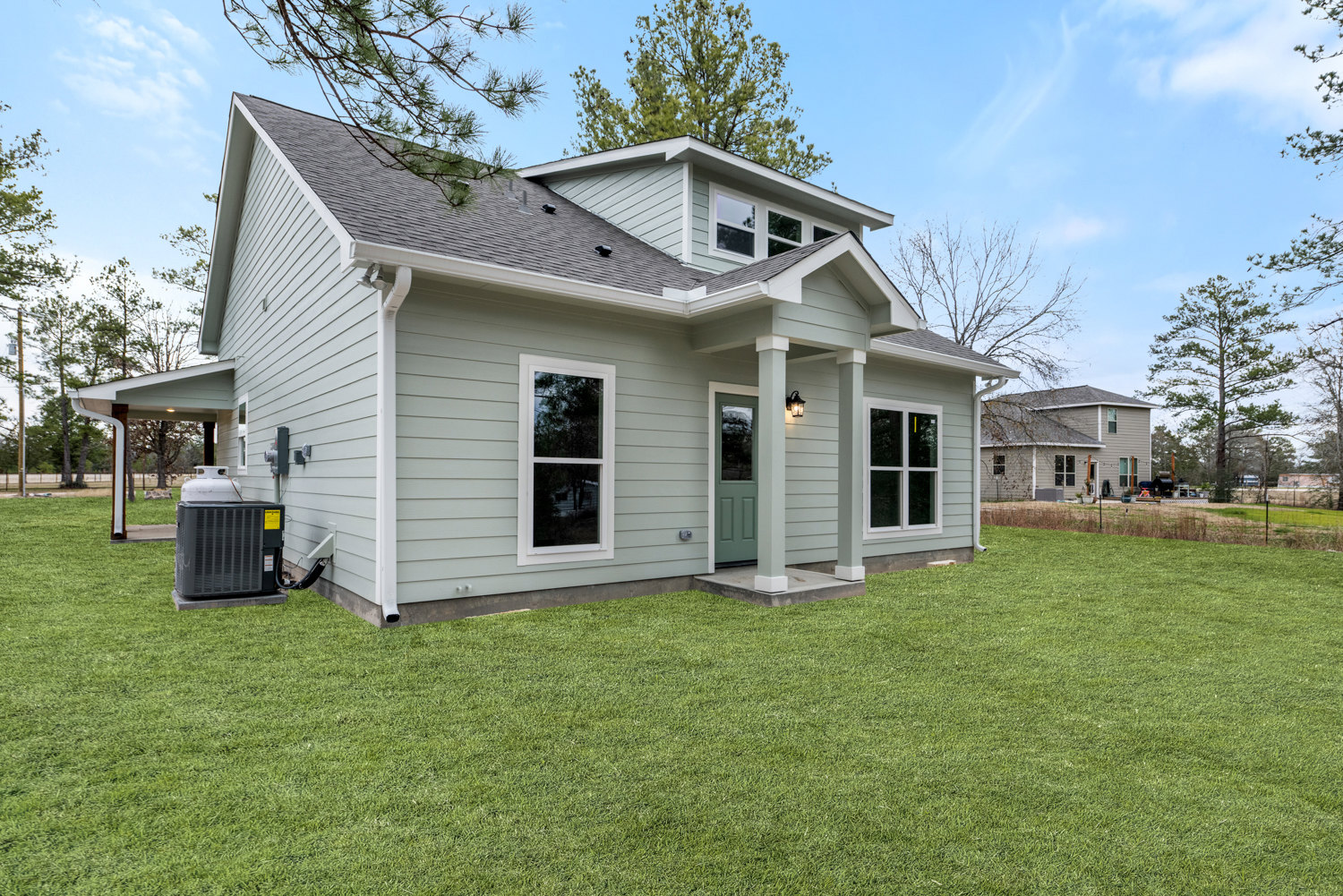 Two-story house with green front lawn, white columns framing the entry, green door, large grey heat pump beside the porch, windows with yellow muntins, mature trees in background