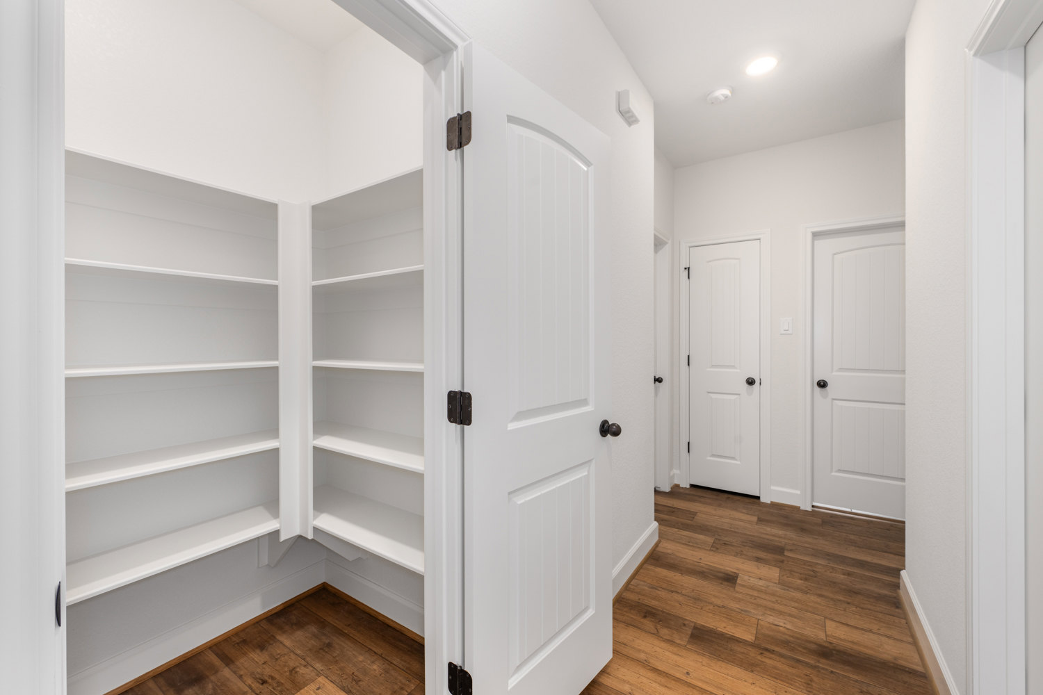 Corner room with white walls, built-in white shelves, hardwood flooring, and a white door featuring a black knob under a ceiling light