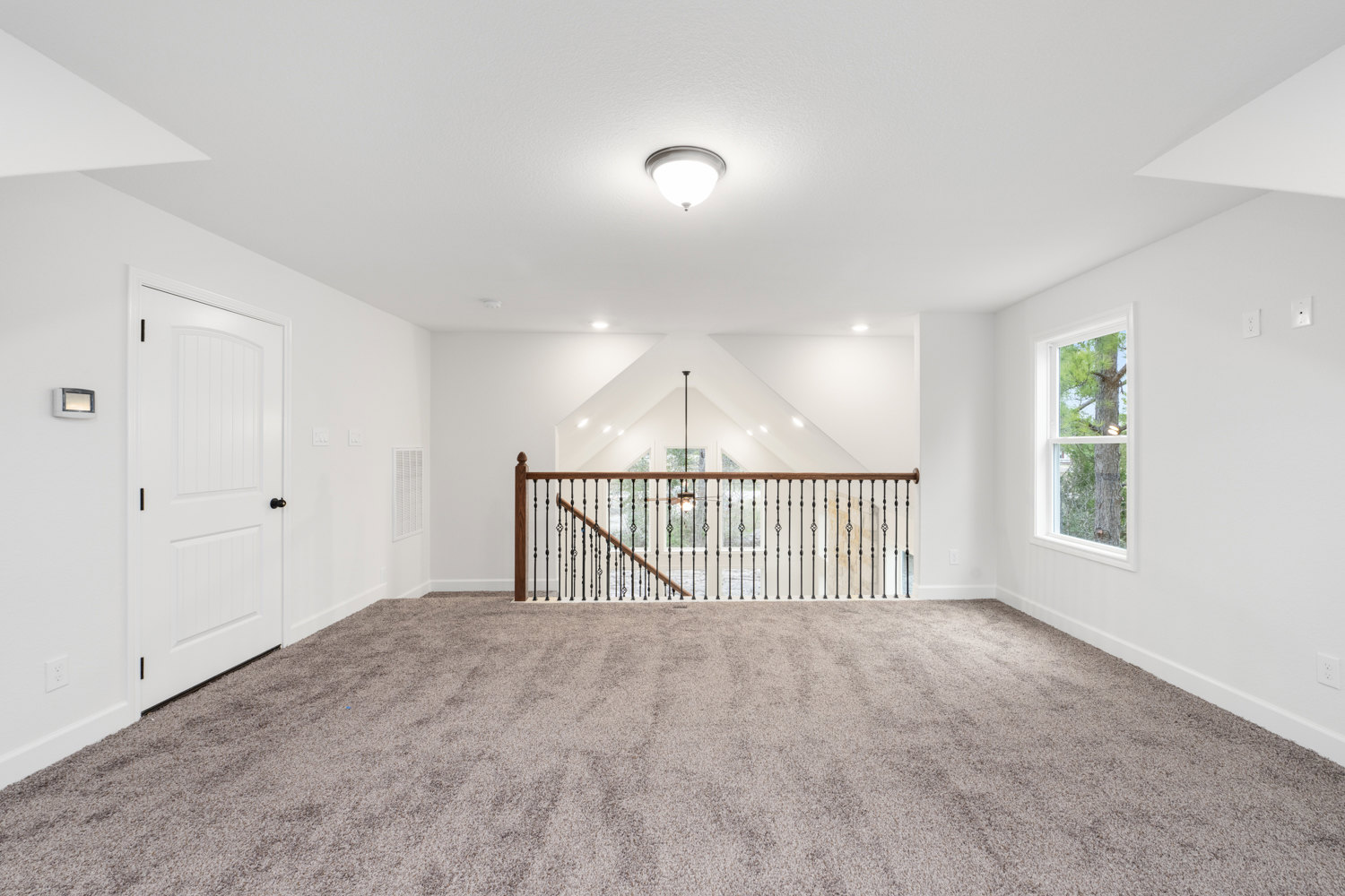 Open living area with carpet flooring, white walls, wooden staircase with black railing, ceiling fan, recessed lighting, white door with black knob, window overlooking leafy tree.