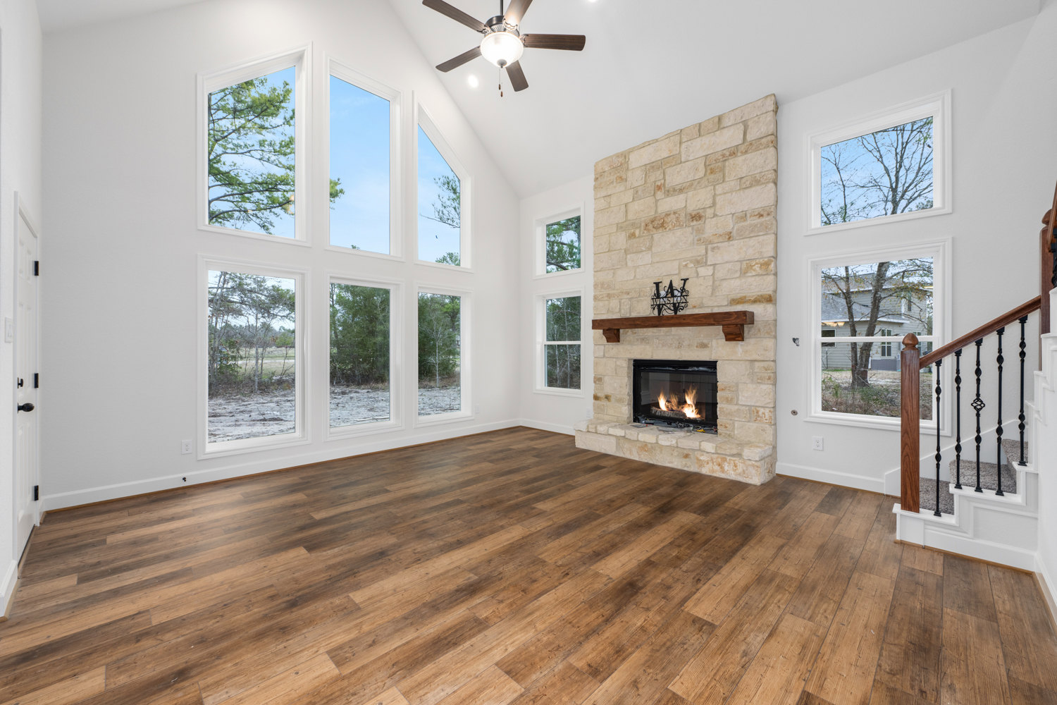 Living room with hardwood floors, stone fireplace with active fire, ceiling fan and light fixture, large window overlooking green trees