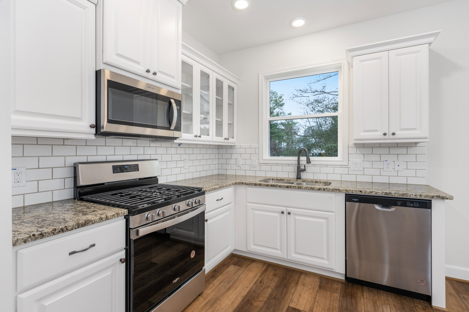 White shaker cabinets, black stove and microwave, stainless dishwasher, quartz countertops, window overlooking trees, white electrical outlet with green indicator light