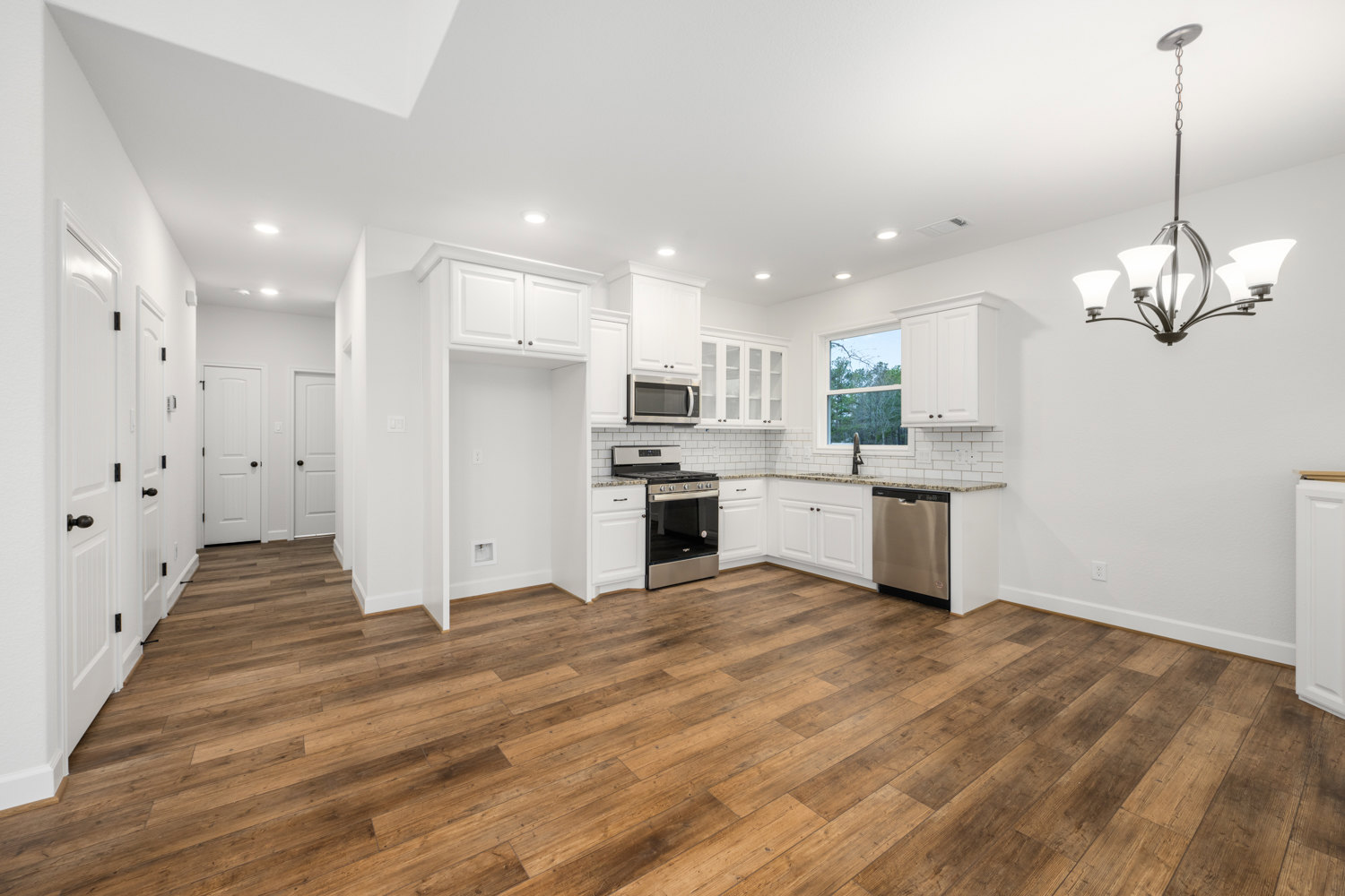 White kitchen featuring wood flooring, white cabinetry, stainless steel stove and oven, open microwave door, close-up of dishwasher, and modern chandelier.