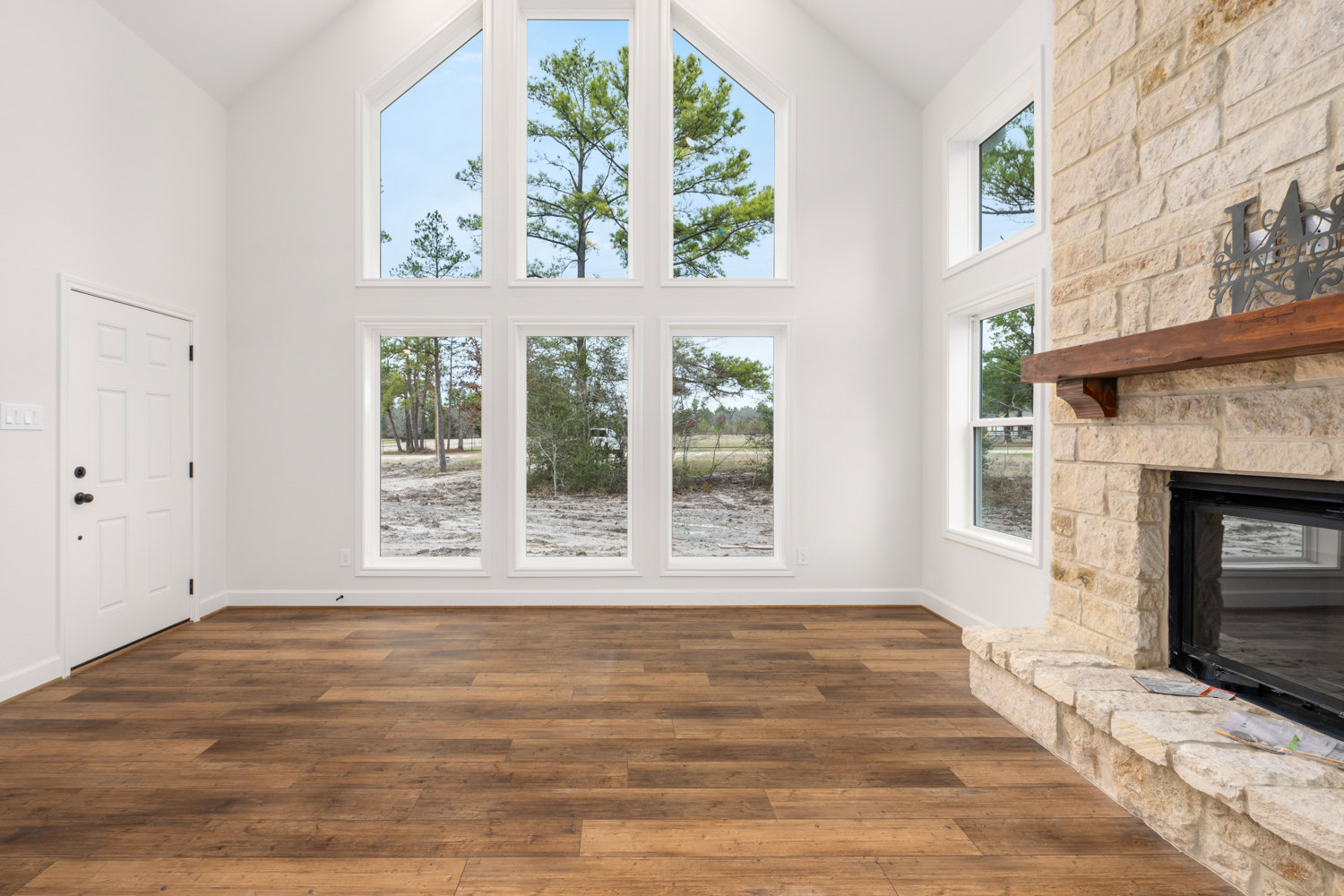 Living room with wide plank wood flooring, white walls, stone fireplace, multiple large windows showing leafy trees outside