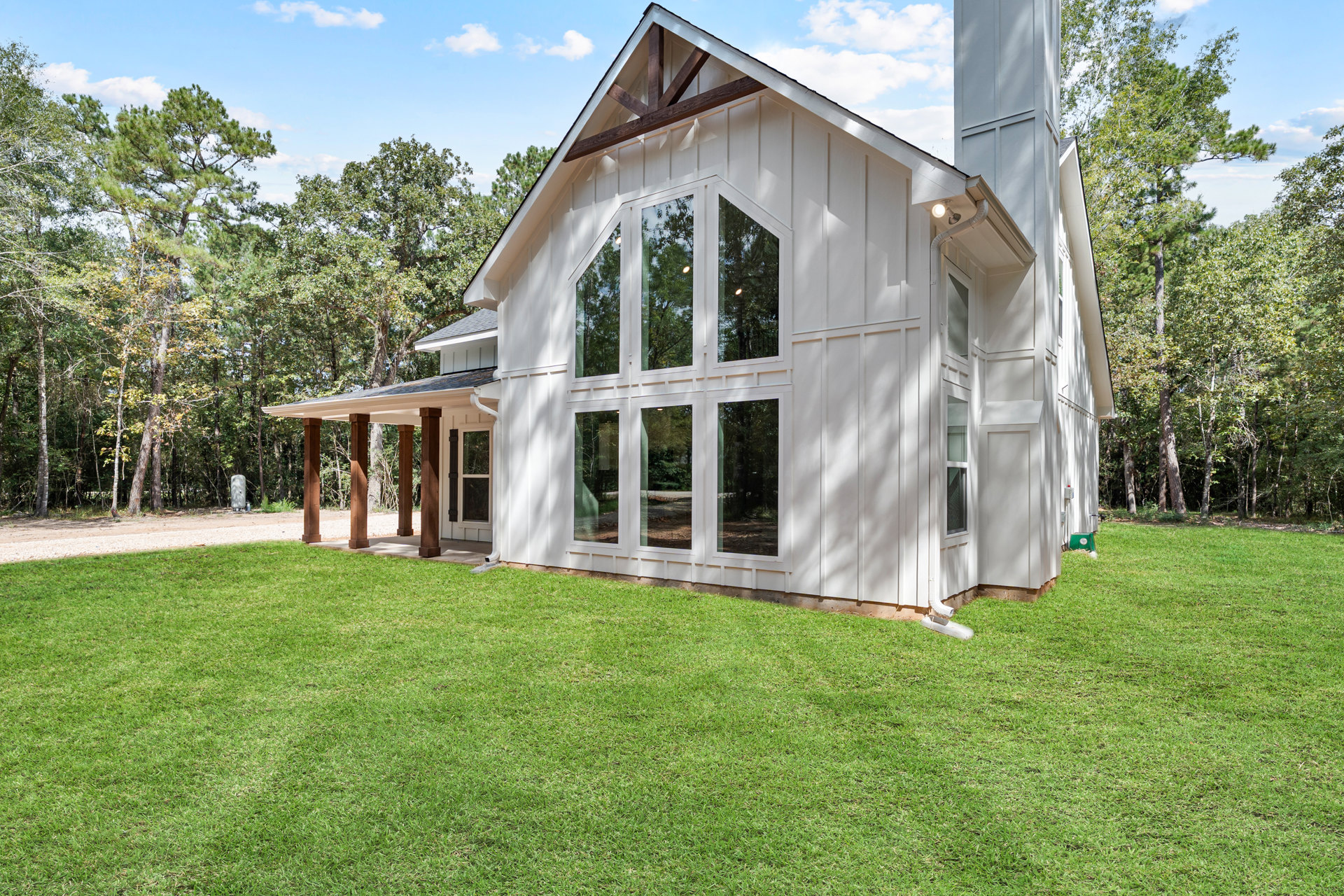 White house with triangular roof, tall brick chimney, green lawn, covered porch with white pillars, trees and cloudy sky in background