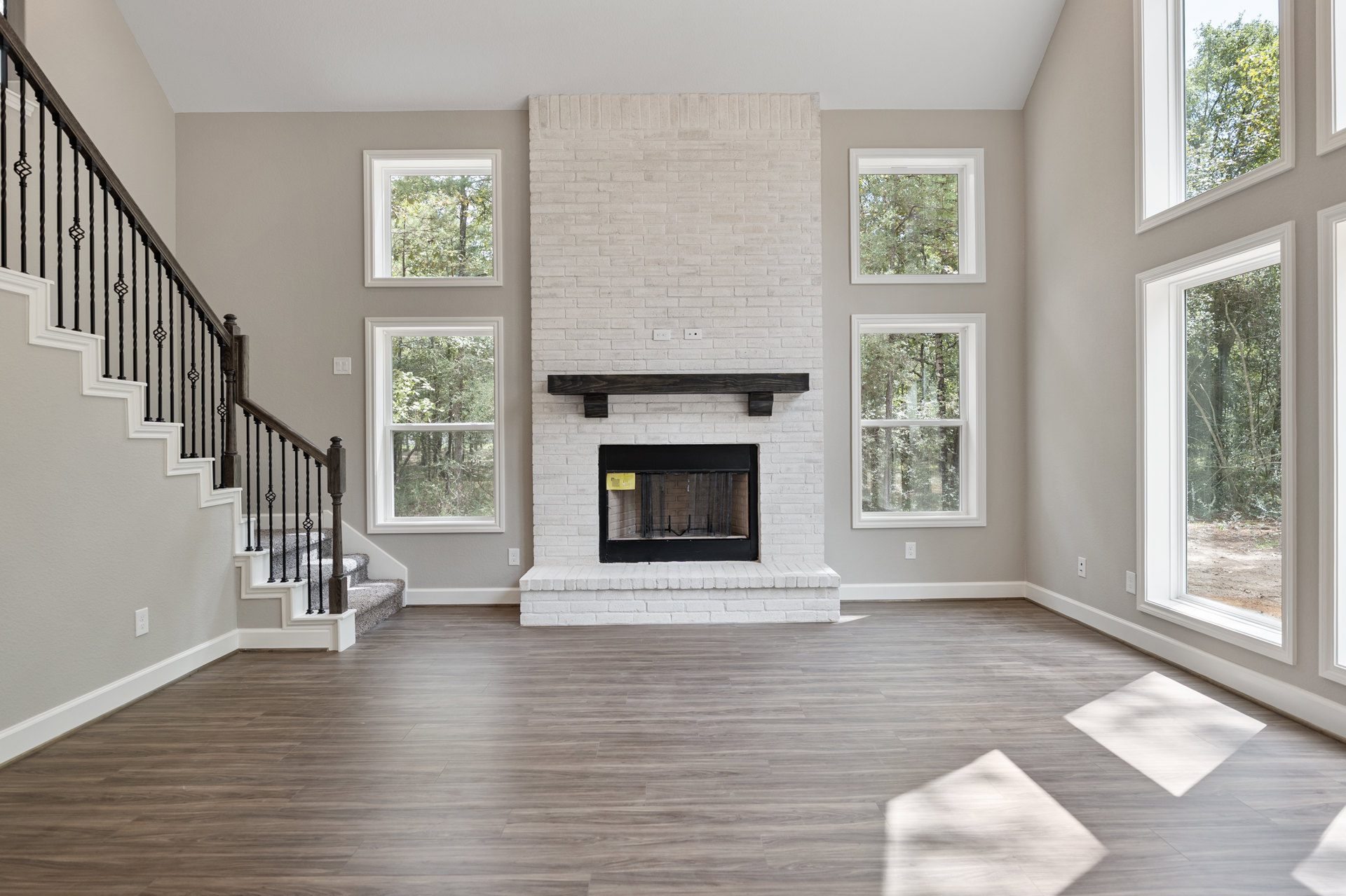 White brick fireplace with black screen, wood flooring, large window revealing trees outside, white walls with minimal molding