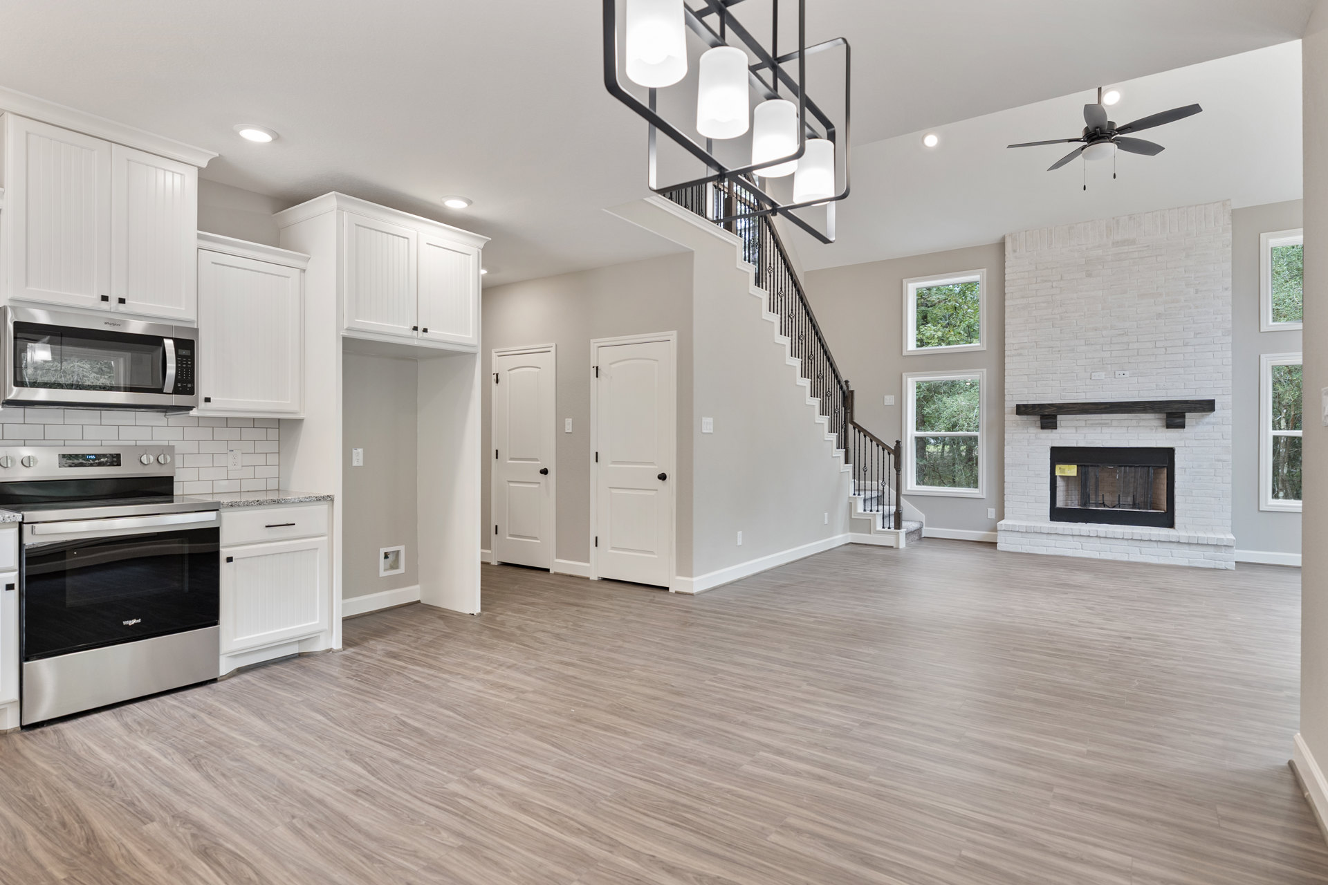 Open living area featuring wood floors, white doors, a staircase with dark railings, stone fireplace, stainless steel oven, built-in microwave, white cabinetry, and ceiling fan
