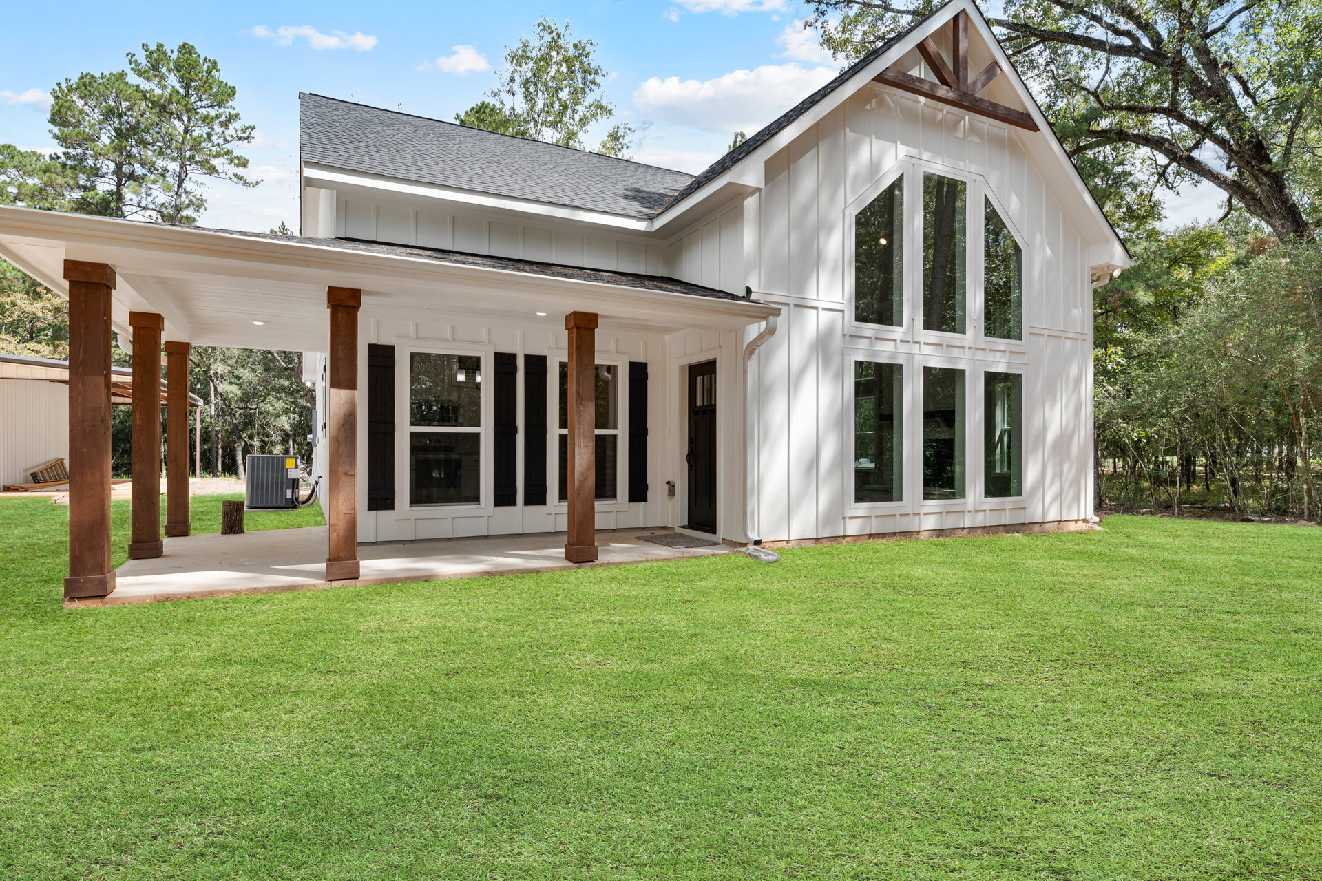 White siding house with black front door, brown porch pillars, large windows, and green lawn bordered by trees.