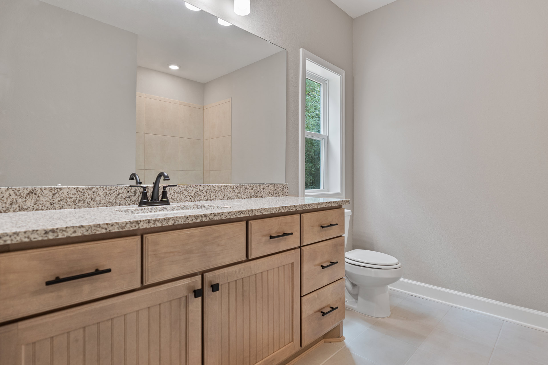 Marble countertop with undermount sink and chrome faucet, white toilet, light gray tile floor, large window overlooking trees, white cabinetry
