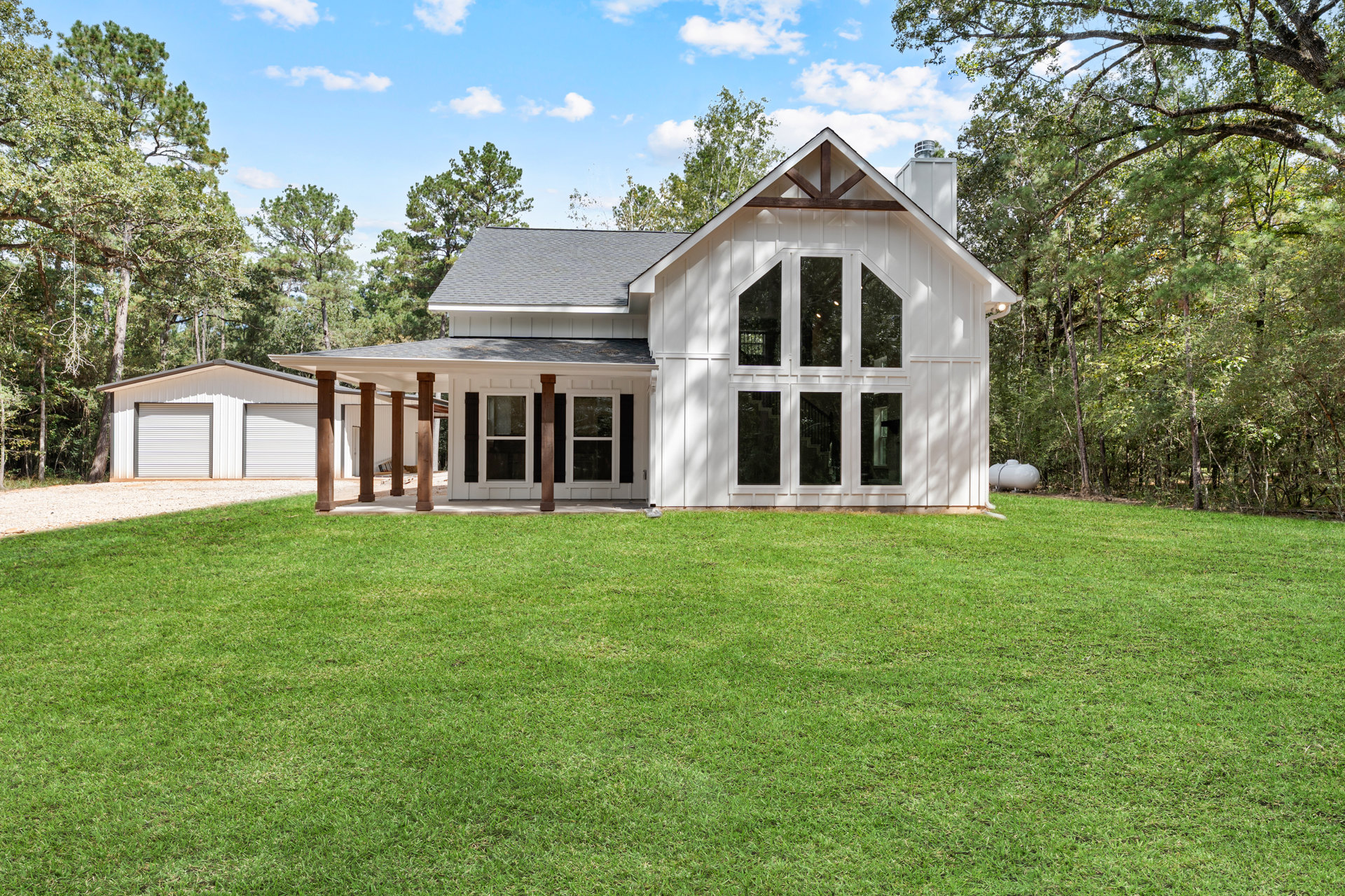 White house with wood roof, large windows, and attached garage door, surrounded by green lawn and trees under partly cloudy sky