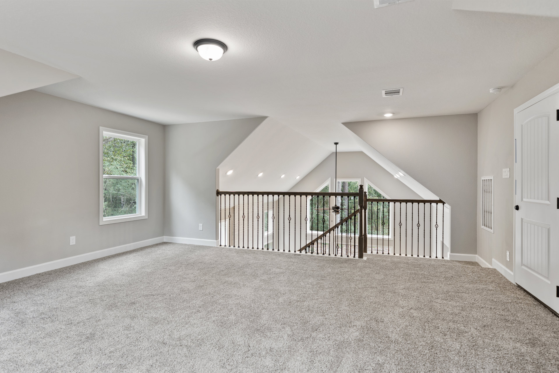 Carpeted room featuring a wooden staircase with black metal railing, large window overlooking trees, white door with black knobs, and ceiling fan.