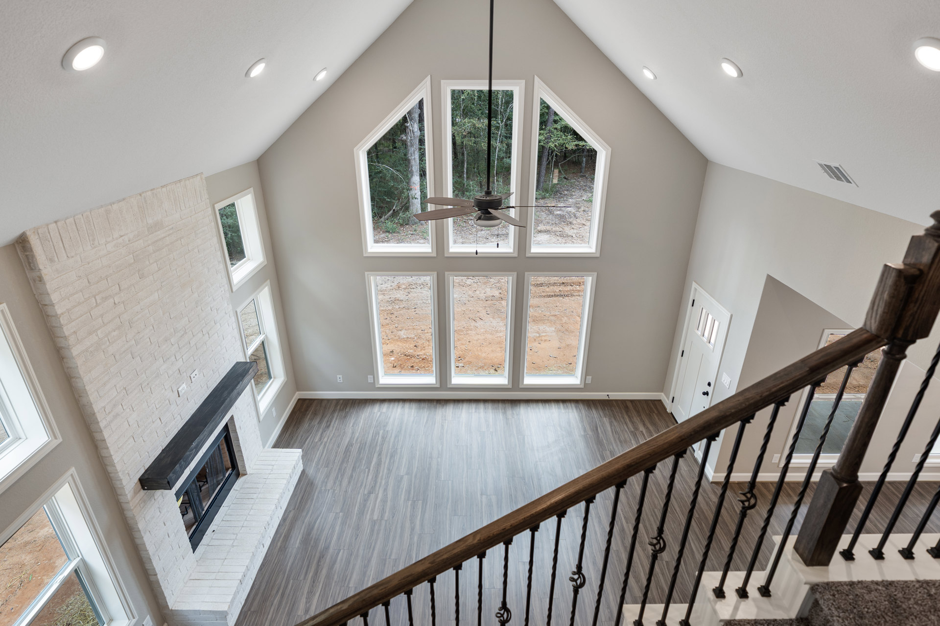 Wooden staircase with white balusters and handrail descending to open living area, stone fireplace, large windows, and modern light fixture