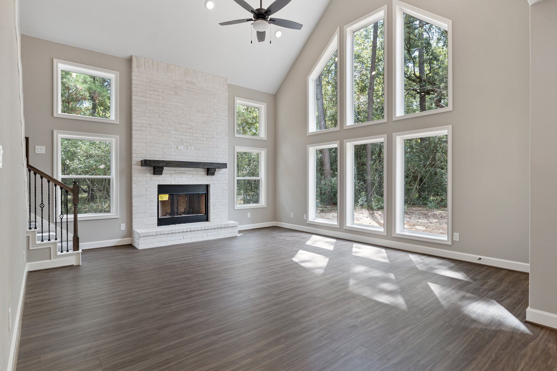 Spacious living area with wood flooring, black-framed fireplace, ceiling fan with light, staircase with metal railing, and large windows overlooking trees