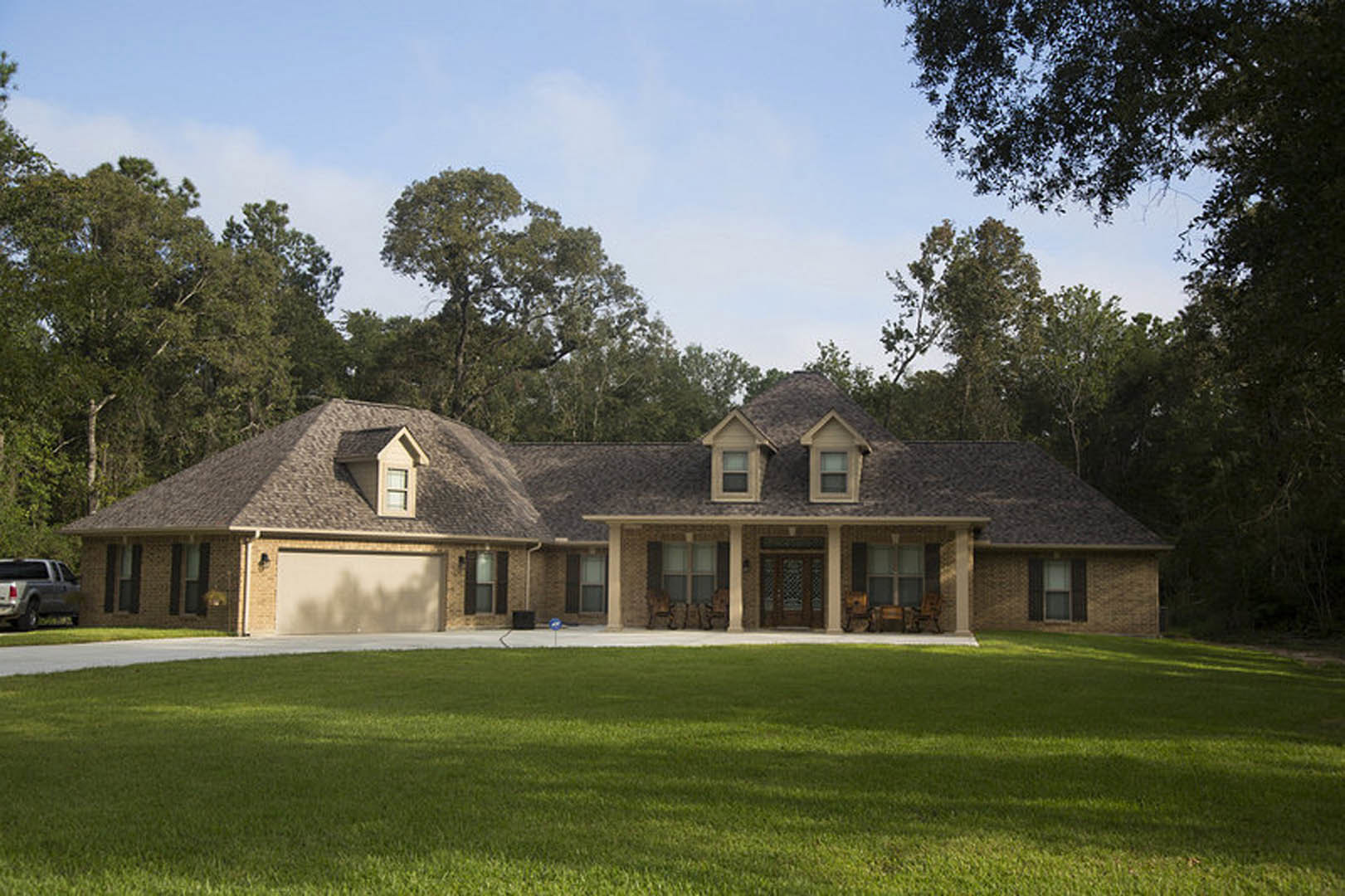 Two-story custom home with white siding, large windows, and a thatched roof, surrounded by a green lawn and mature trees under a clear sky