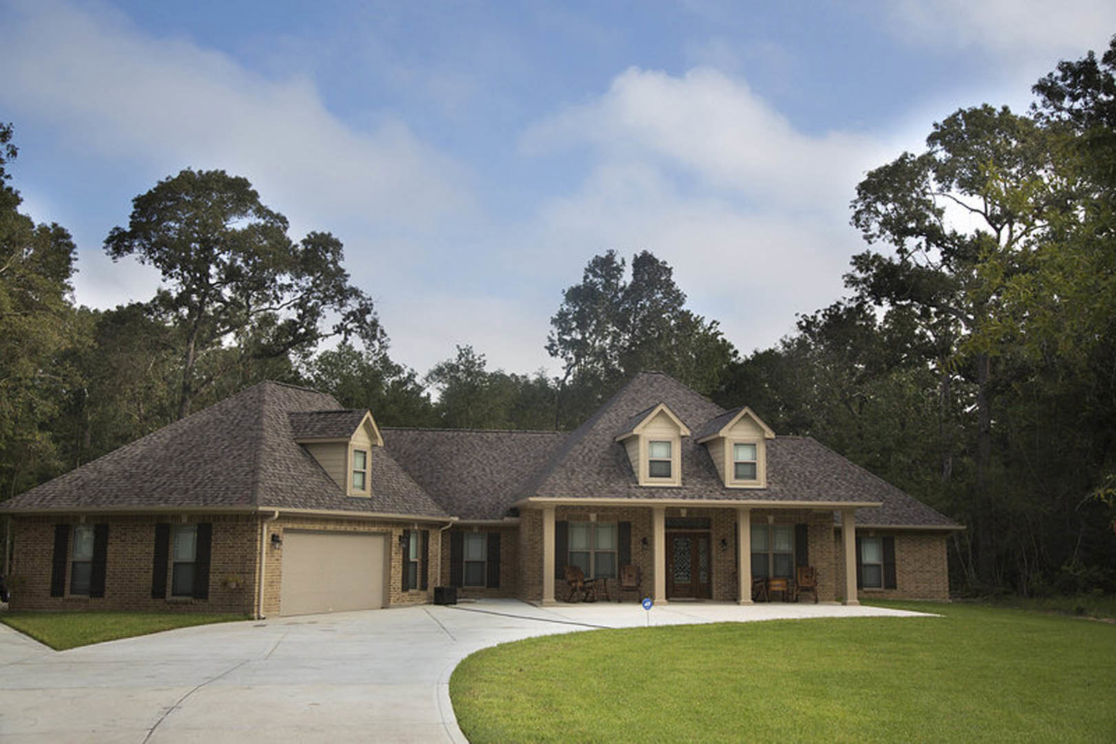 Two-story house with gray siding, attached garage, covered front porch, concrete driveway, and manicured green lawn bordered by trees.