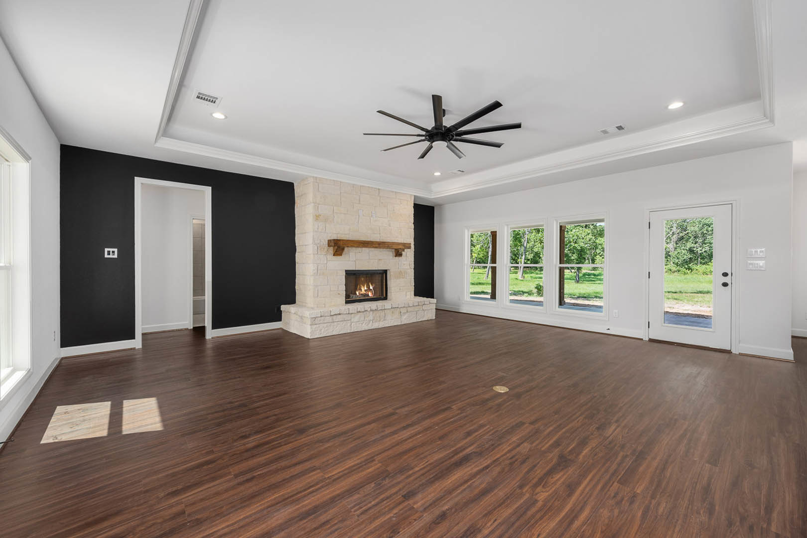 Spacious living area featuring hardwood floors, stone fireplace with active fire, ceiling fan with light, large windows, and glass door overlooking grassy yard and trees.