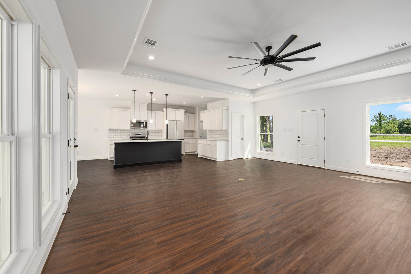 Open-concept kitchen with wood flooring, black accent wall with white trim, ceiling fan with light, white door with black hardware, and large windows showing blue sky and trees