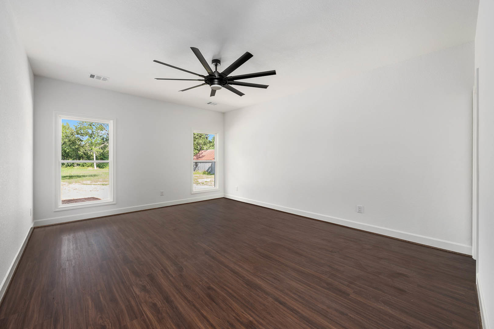 Ceiling fan with light fixture, dark hardwood floor, white plaster walls, large window overlooking trees, red roof, and fenced grassy yard