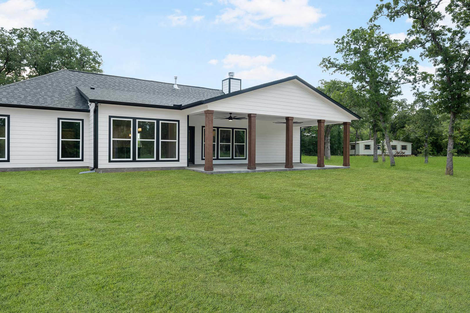 White house with black trim, brown pillars, and white door, set on a green lawn; chimney on roof, blue sky and trees in background.