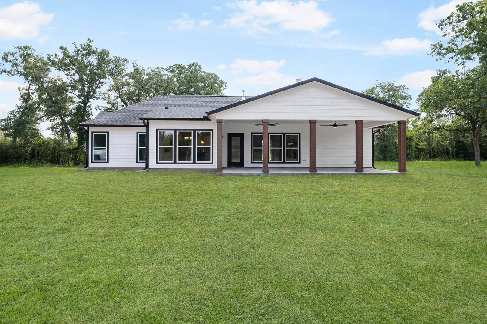 White house with black trim, covered front porch supported by columns, manicured green lawn, large windows with white frames, trees and plants bordering the property, partly cloudy