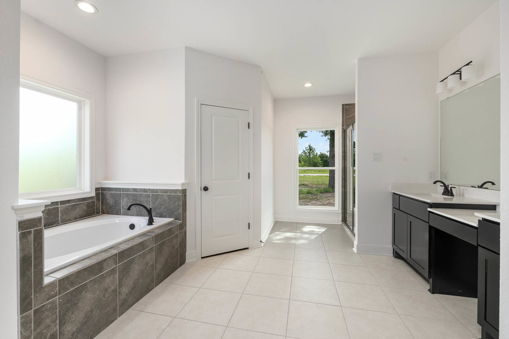 Bathroom with freestanding tub against a tiled wall, white tile floor, white door with black knobs, single sink vanity, window overlooking tree and grass.