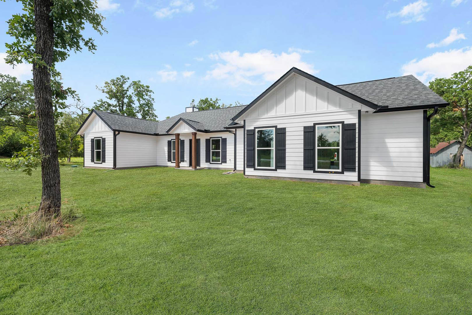 White two-story house with black shutters, white-framed windows, and a green lawn bordered by mature trees under a partly cloudy sky