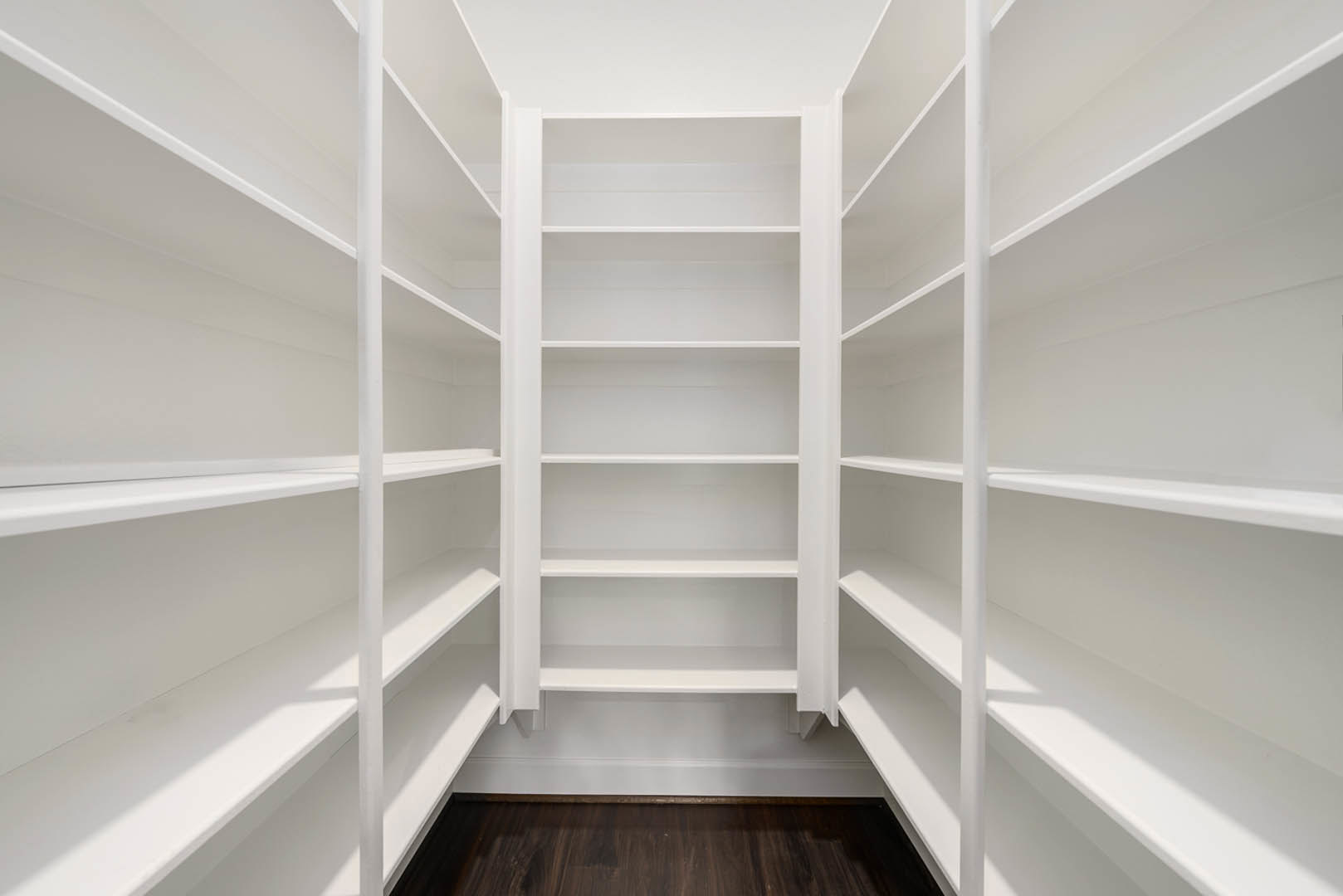 White built-in shelving unit against a white plaster wall, wood floor visible, white ceiling above, minimalist pantry interior.