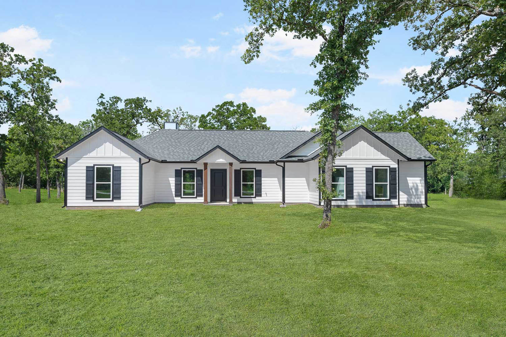 White two-story house with black front door, white-framed windows, manicured green lawn, and leafy tree in front yard under partly cloudy sky
