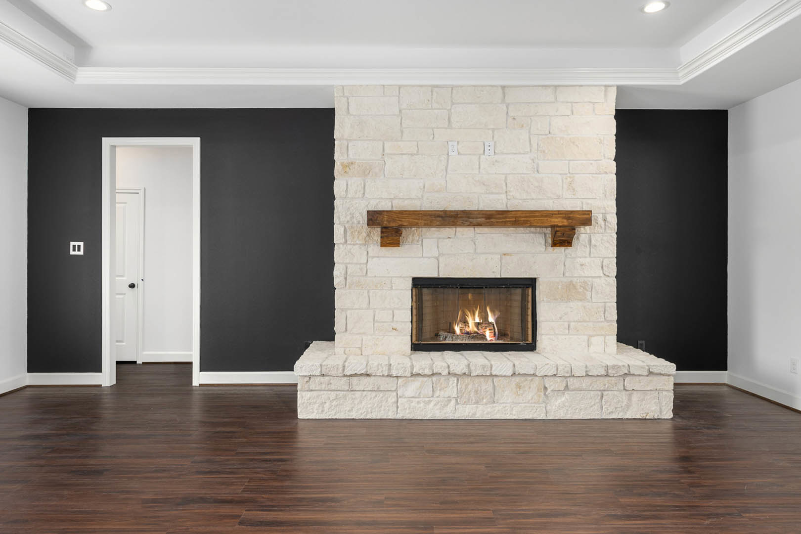 Stone fireplace with active fire, black metal fire screen, wood sign mounted on brick wall, white door with black knobs, hardwood flooring, neutral walls