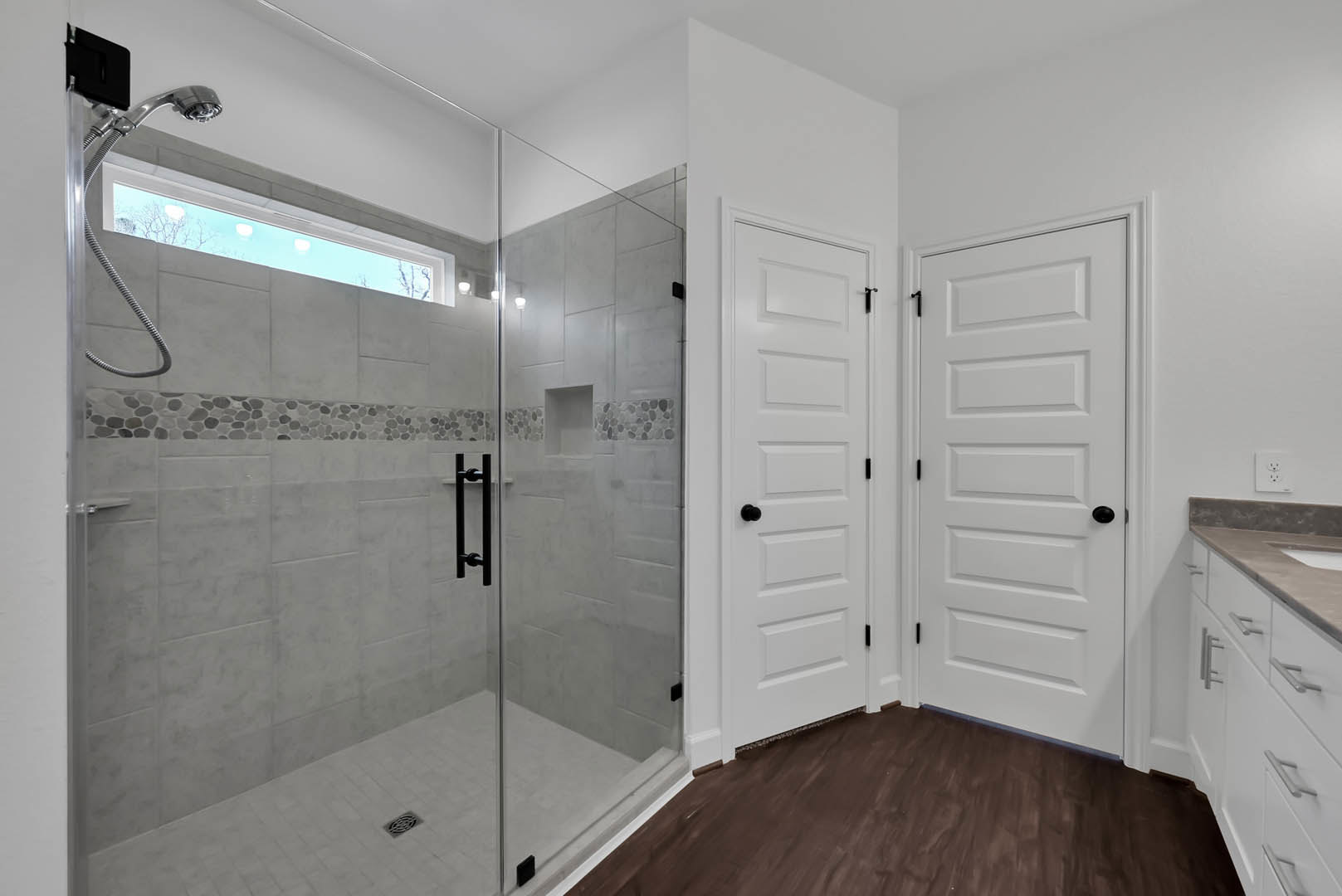 Bathroom with glass shower enclosure, white door with black knob, dark wood flooring, white walls, window, and white electrical outlet.