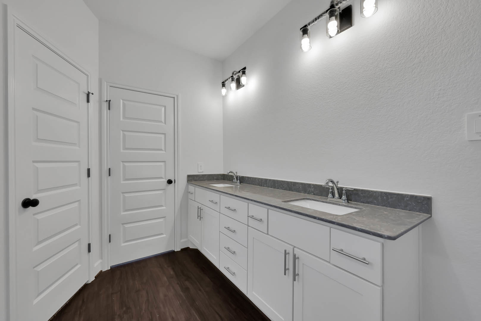 Bathroom featuring white shaker cabinets, dark wood flooring, white door with black hardware, wall-mounted light fixture, and quartz countertop with chrome faucet.