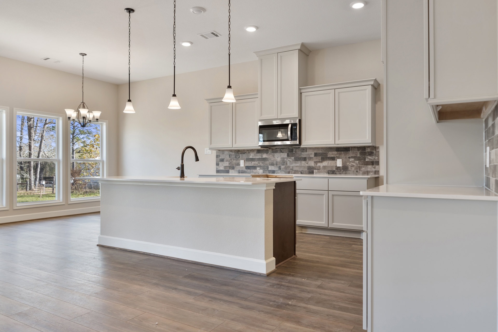 Open kitchen featuring a central island with white countertop, wood flooring, white cabinetry, built-in microwave near window, decorative vase on island, and modern pendant light