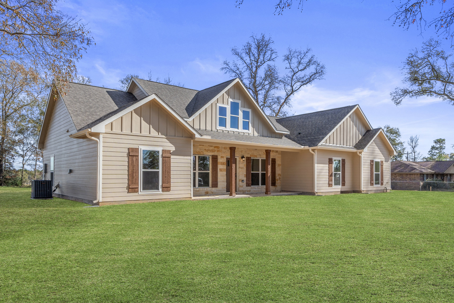 Modern stone house with white-framed windows, covered porch supported by columns, manicured green lawn, and mature trees under a clear sky