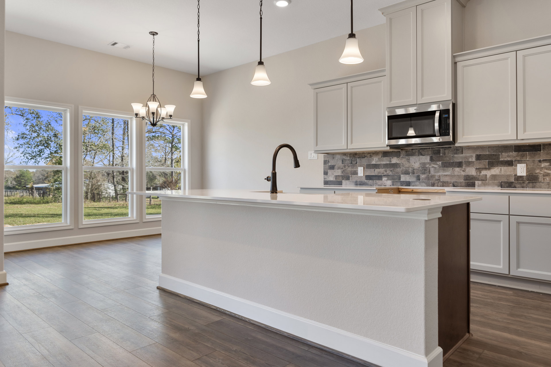 Spacious kitchen featuring a large central island with granite countertop, wood flooring, white cabinetry, stainless steel microwave, modern faucet, and pendant lighting fixtures