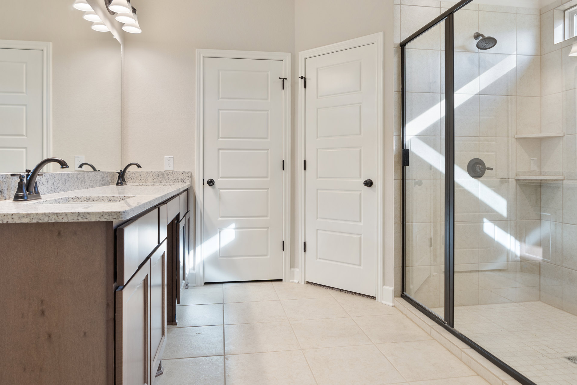 Bathroom featuring white paneled doors with black handles, glass-enclosed shower, white tile walls, modern sink with chrome faucet, and overhead light fixtures.