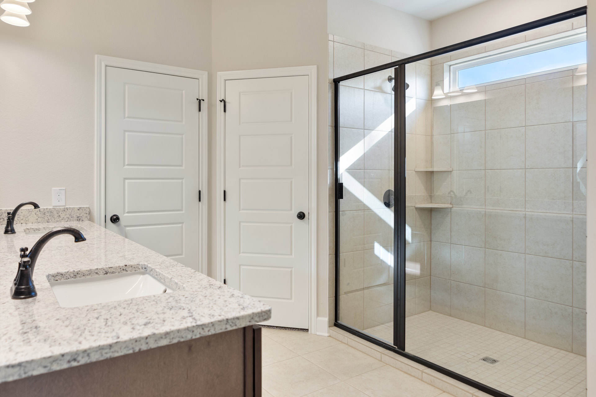 Bathroom with glass shower enclosure, white sink set in stone countertop, black faucet, white door with black hardware, light tile walls, and modern fixtures.