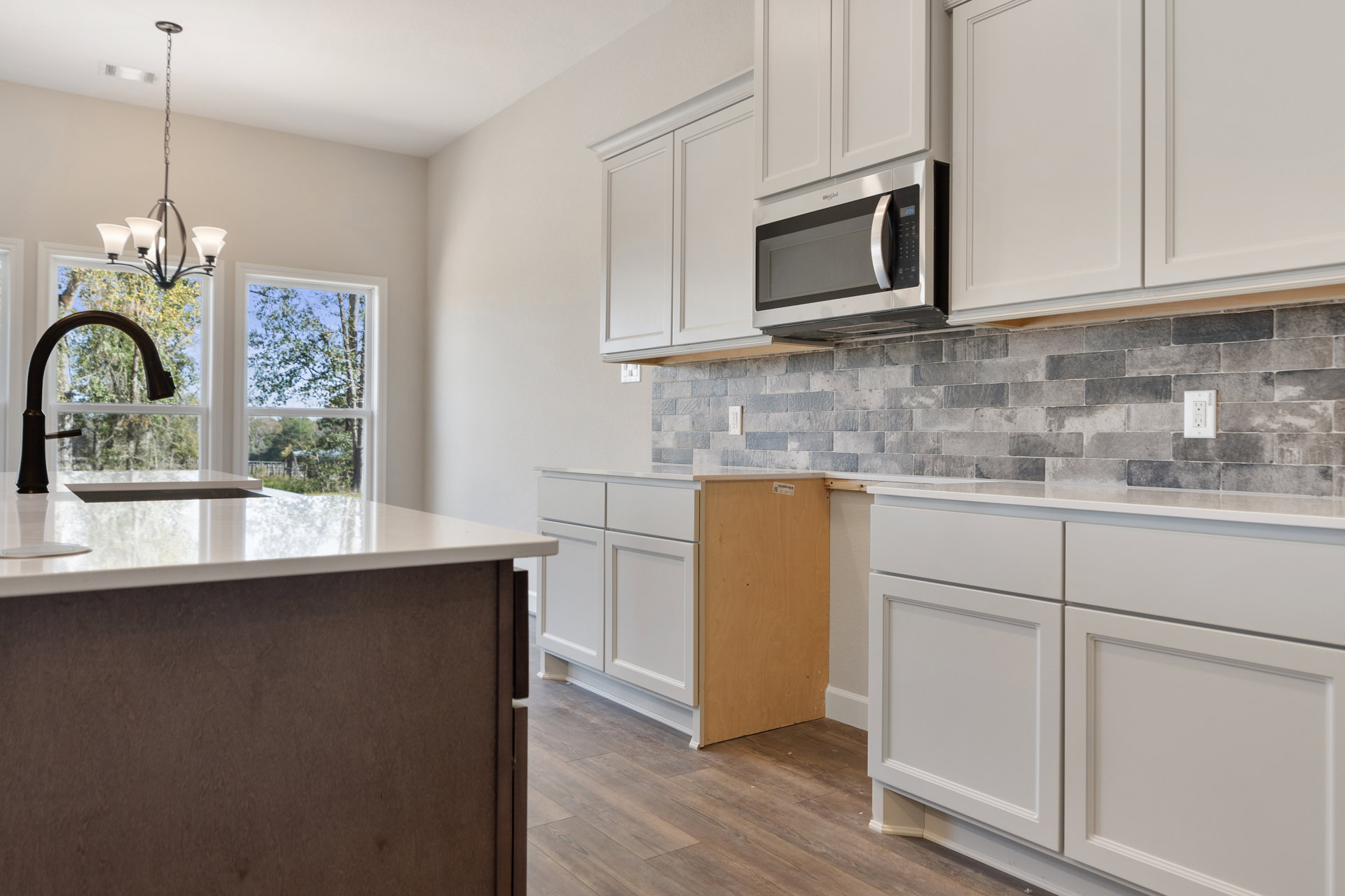 White kitchen cabinets with grey tile backsplash, granite countertops, stainless steel microwave, wood flooring, and a sink with a faucet set beneath a window.