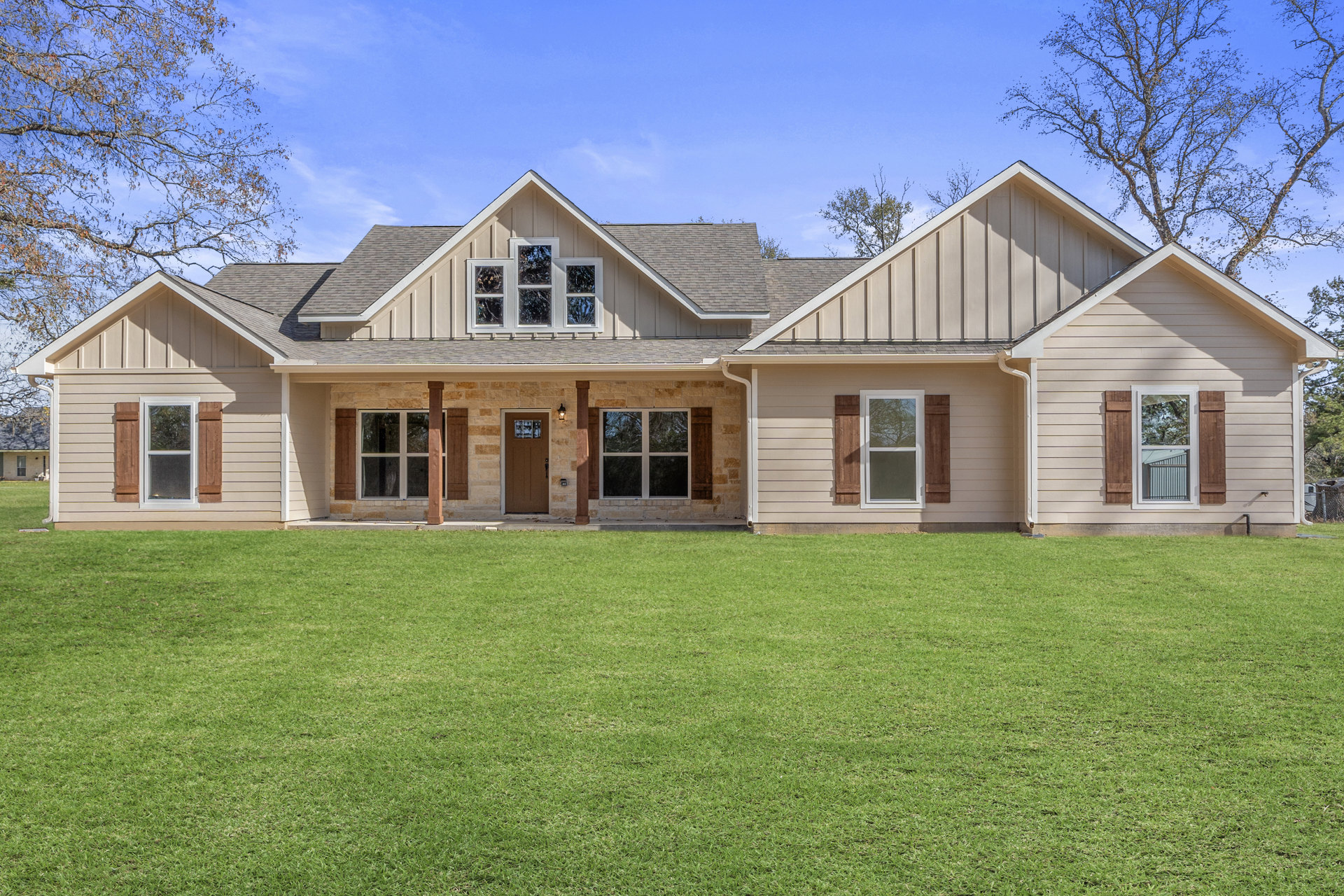 Stone exterior custom home with shingle roof, white-framed windows reflecting trees, green lawn, and close-up of wood door.